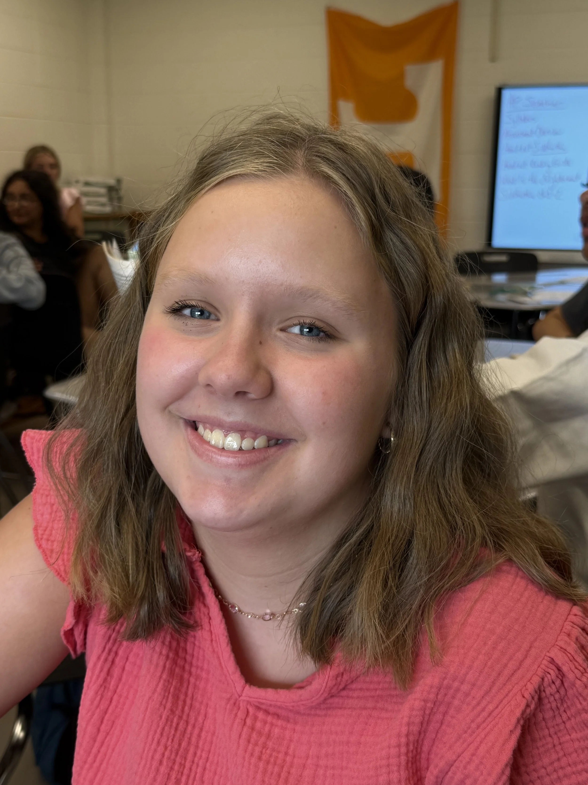 A young girl with braided hair wearing a green hoodie, sitting at a table in a classroom or meeting room, with a whiteboard and two other students in the background.