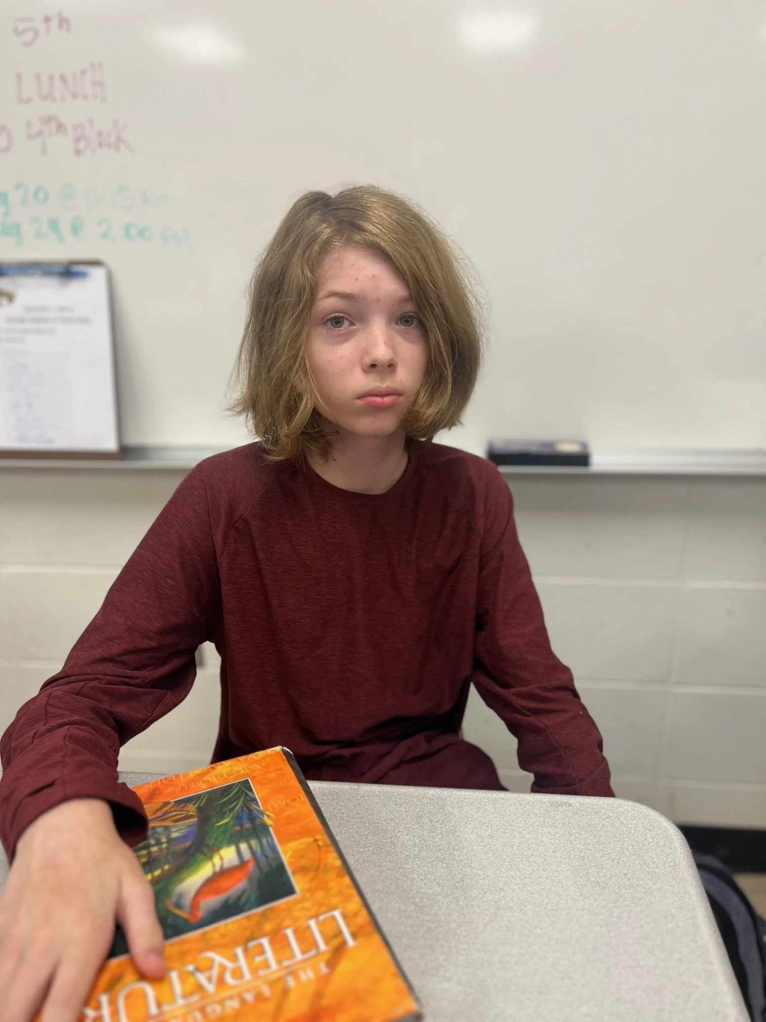 A young woman with long wavy hair and freckles, smiling at the camera, sitting in a classroom with a whiteboard and shelves of binders behind her.