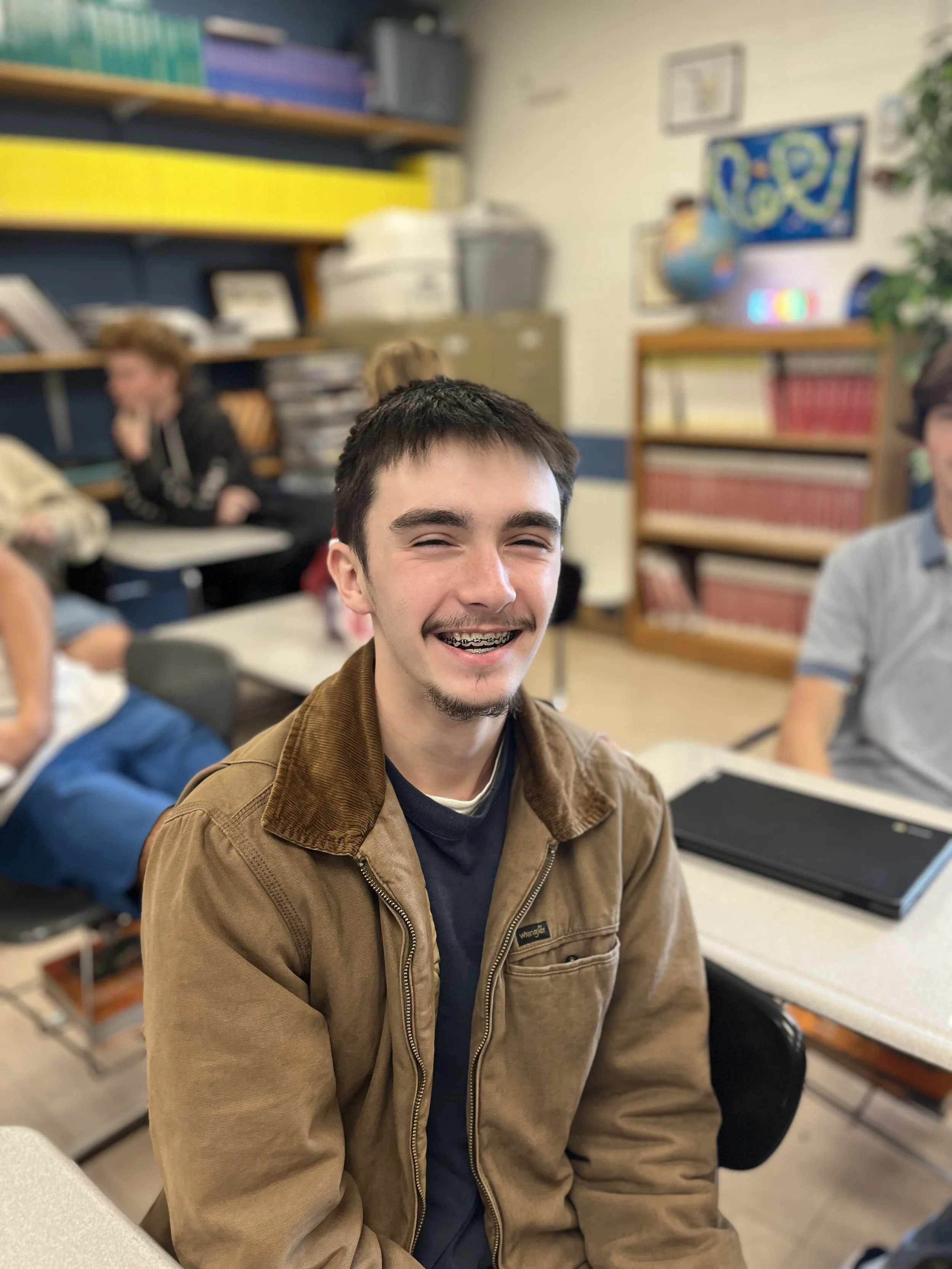 A young man smiling, wearing braces and a gray hoodie, sitting indoors with a blurred background that includes a yellow wall and some posters.