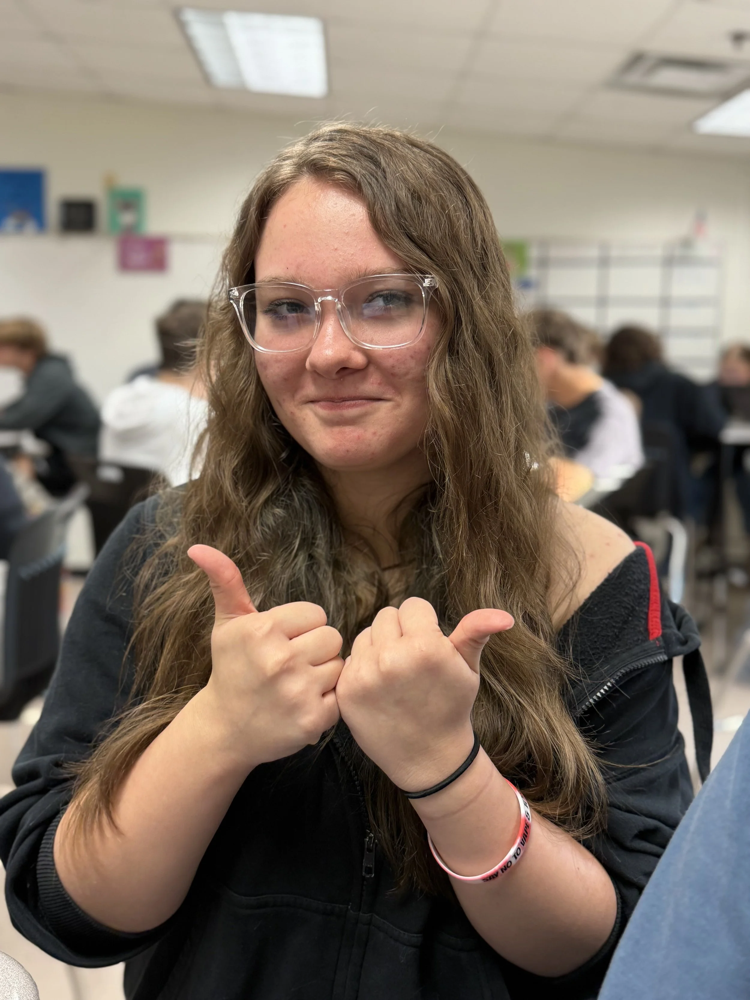 A teenage girl with long wavy brown hair, wearing glasses, smiling and giving two thumbs up in a classroom with other students in the background.