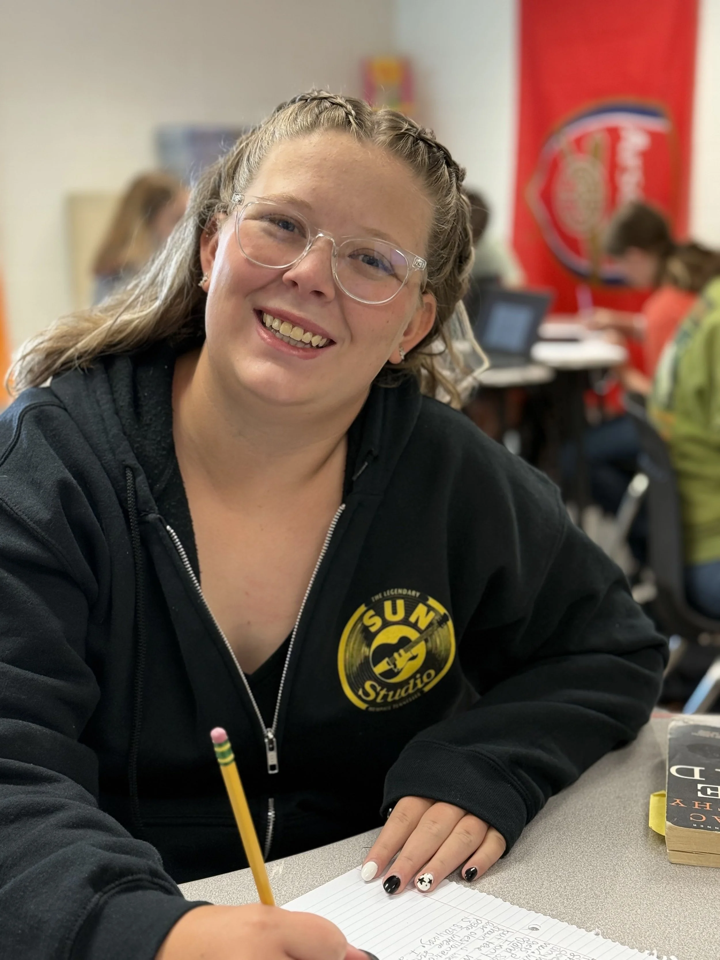 A smiling woman with glasses and braided hair wearing a black hoodie with a yellow 'Sun Studio' logo, sitting at a desk with a yellow pencil and notebook, in a classroom setting with other students and a red wall in the background.