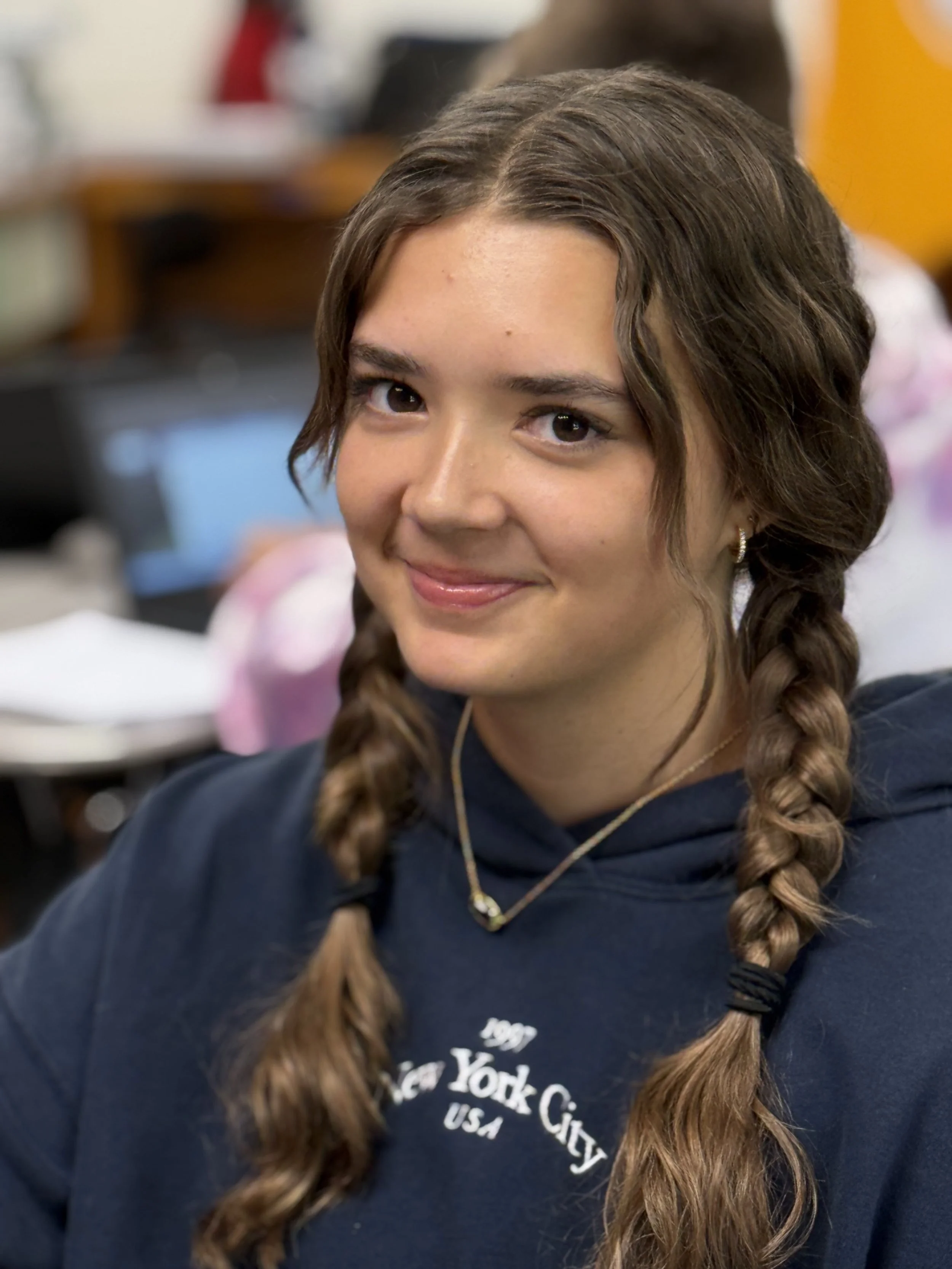 A young woman with long braided hair, wearing a navy hoodie and a gold necklace, smiling at the camera in a classroom or casual setting.