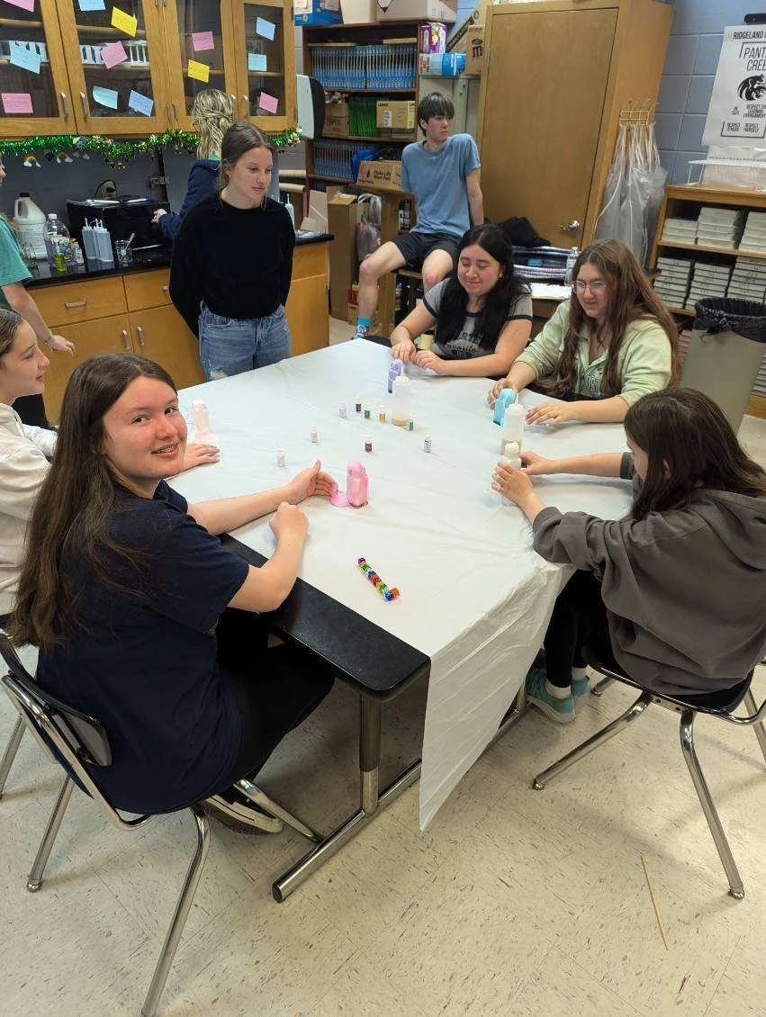Group of students sitting around a table in a classroom, engaging in a hands-on science activity. Some students are holding small bottles and looking at colorful beakers, while others watch and participate. Classroom shelves and cabinets are visible 