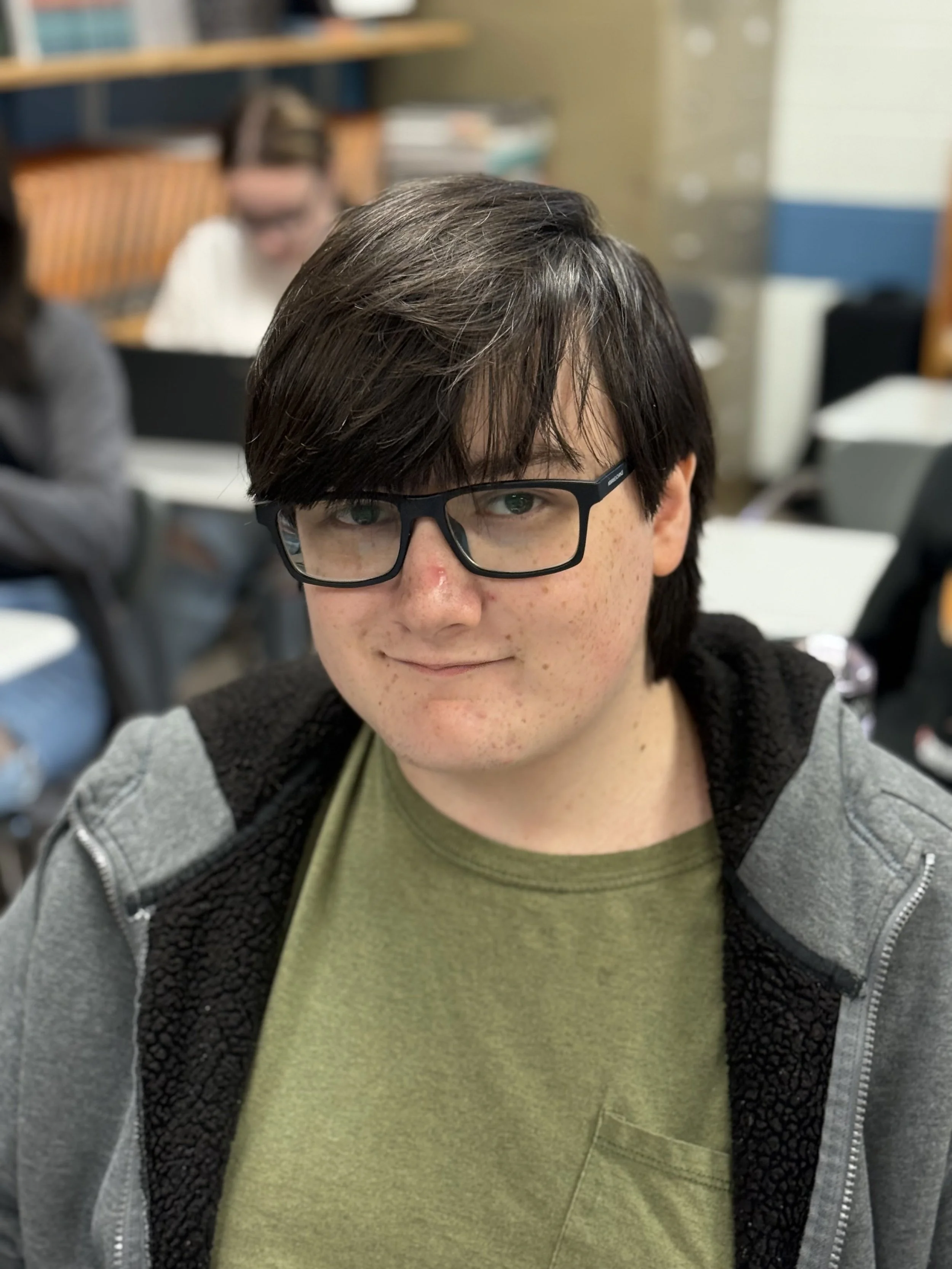 A young man with dark hair, glasses, and light skin, smiling in a classroom or library setting. He's wearing a green shirt and a grey zip-up hoodie. There are other people and books in the background.
