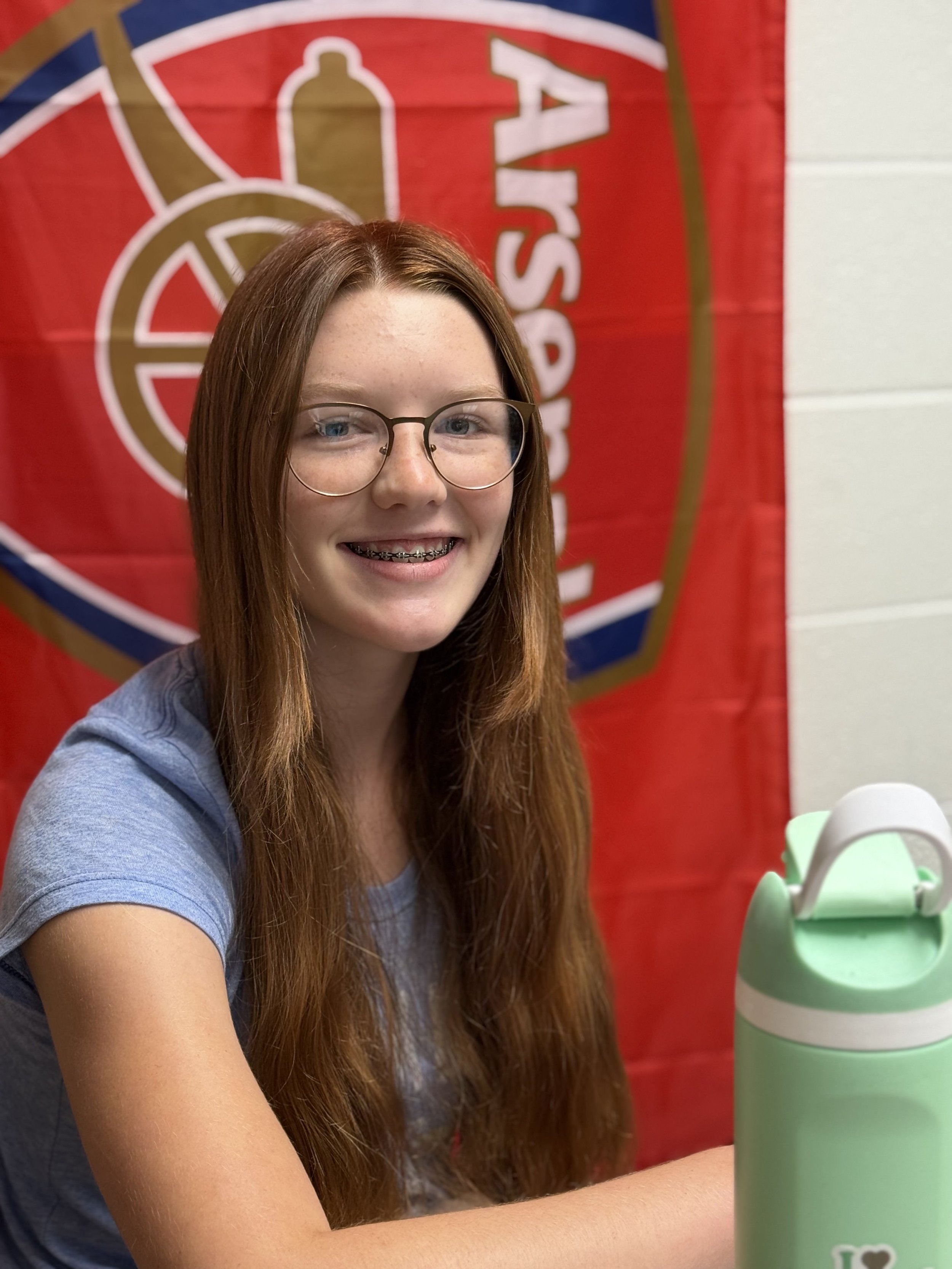 A young woman with long red hair, glasses, and braces smiling while sitting at a table in front of a red banner with a beige and gold emblem. There is a light green water bottle on the table.