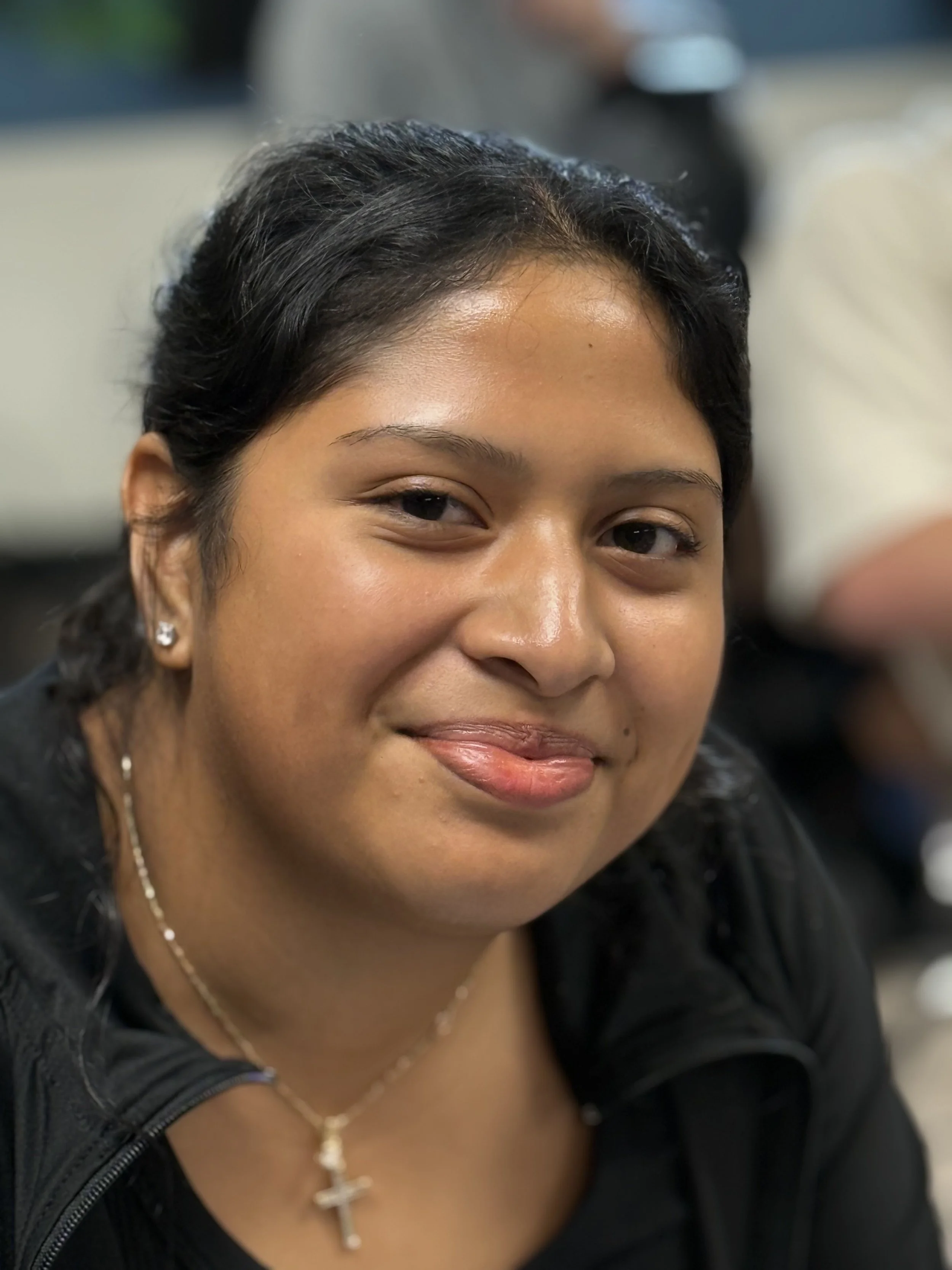 Close-up of a woman with dark hair, wearing earrings and a necklace with a cross pendant, smiling slightly, in an indoor setting.