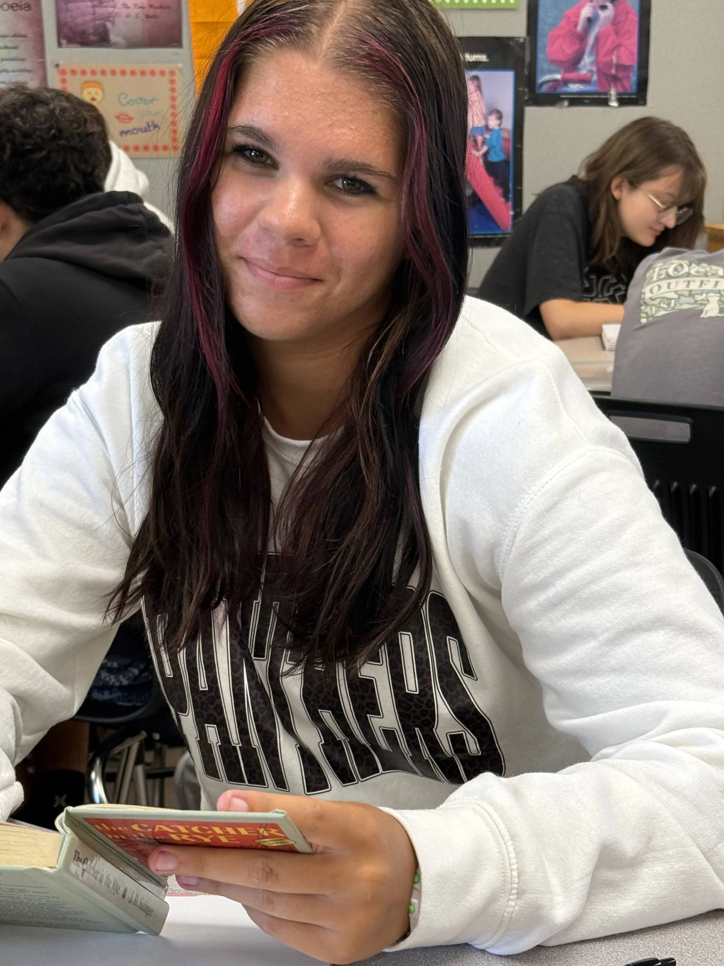 Teen girl with brown hair and a black sweatshirt sitting at a school desk in a classroom, looking at the camera, with other students working on laptops around her.