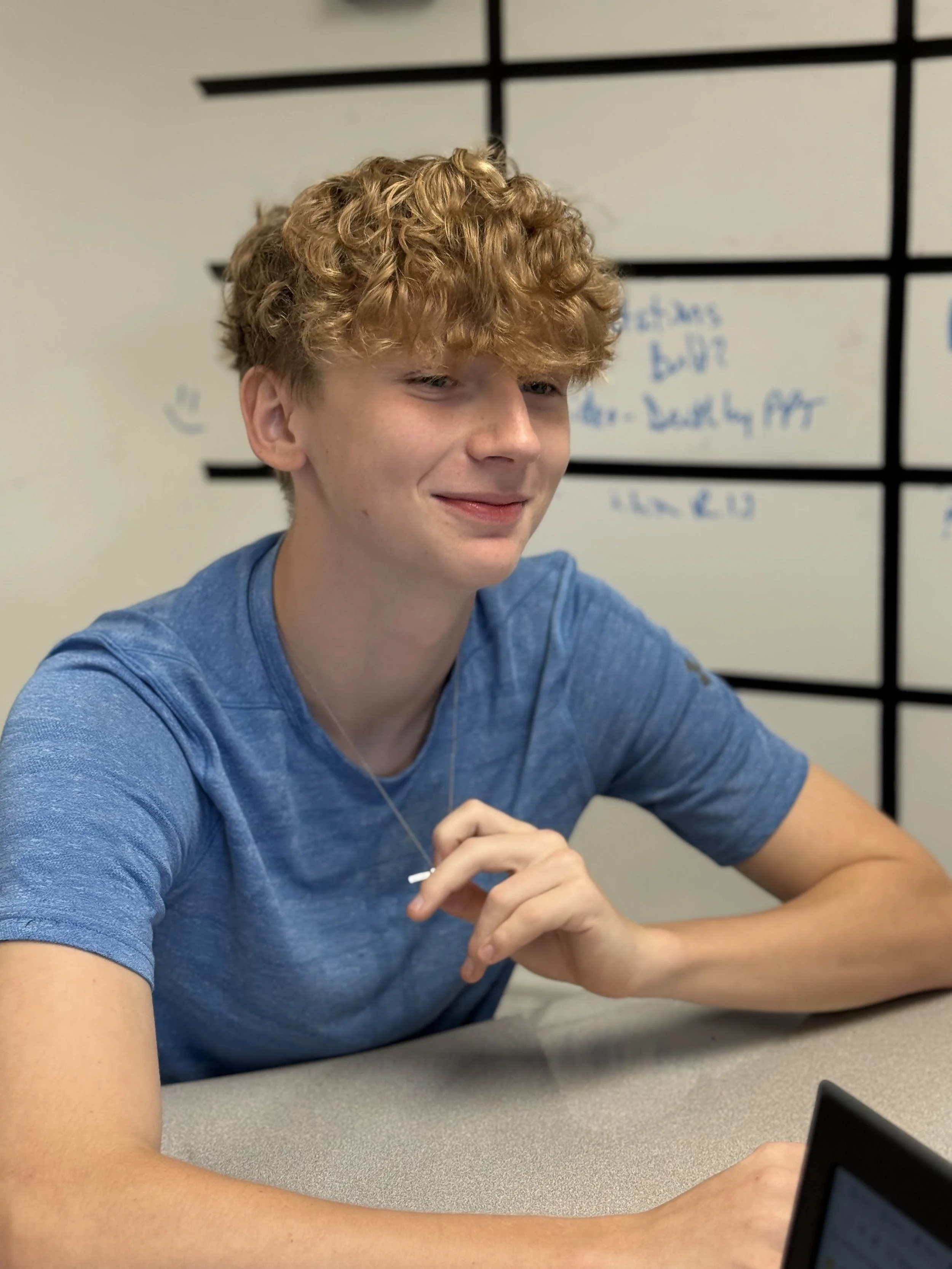 A young man with curly red hair sitting at a table in a classroom or meeting room. He is wearing a blue T-shirt and a silver necklace, smiling slightly, with a whiteboard in the background.