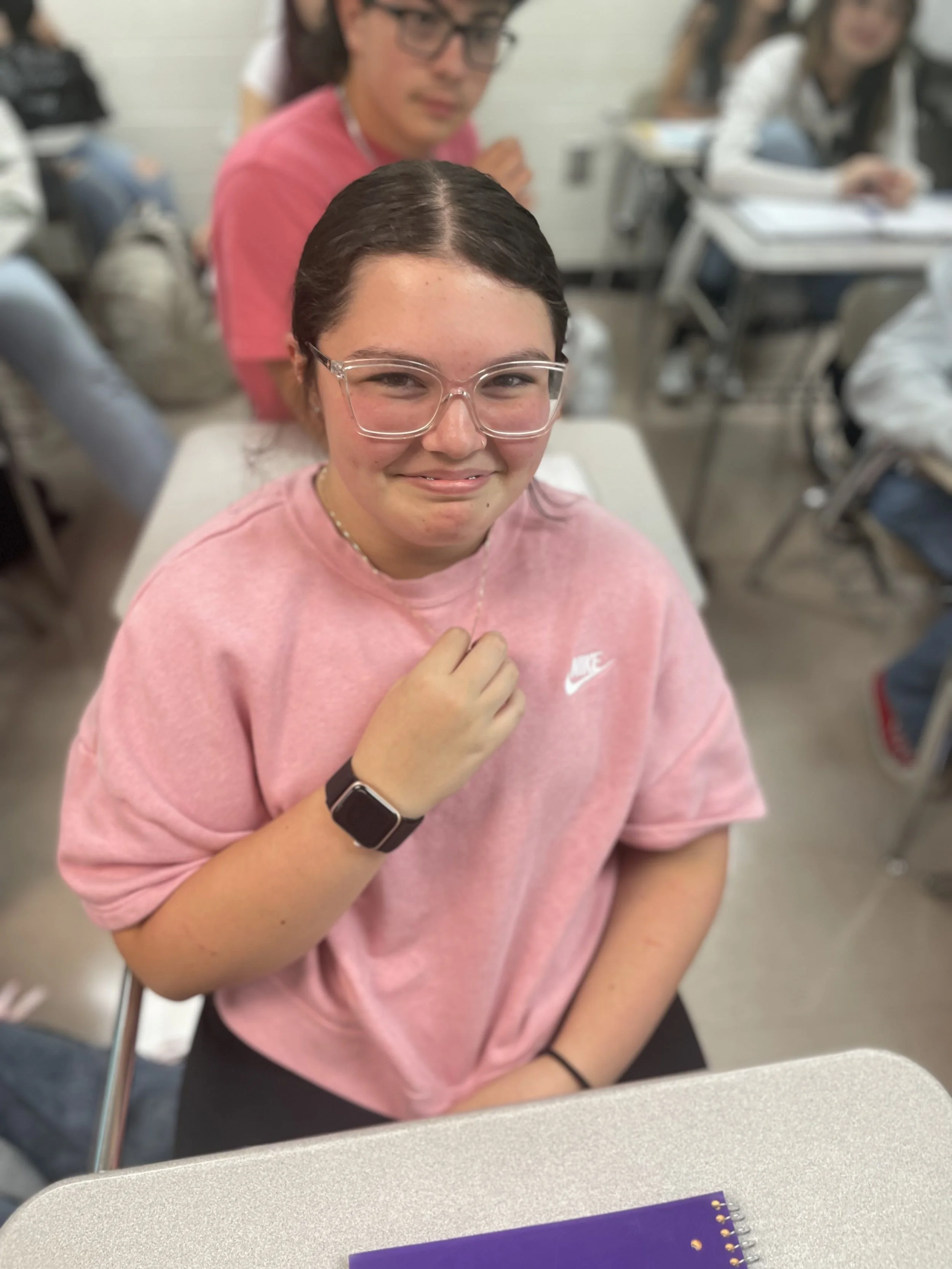 A girl with glasses and a pink Nike t-shirt is sitting at a desk in a classroom, smiling and looking at the camera. She is wearing an Apple Watch and a necklace, with a purple notebook in front of her. Other students are visible in the background.