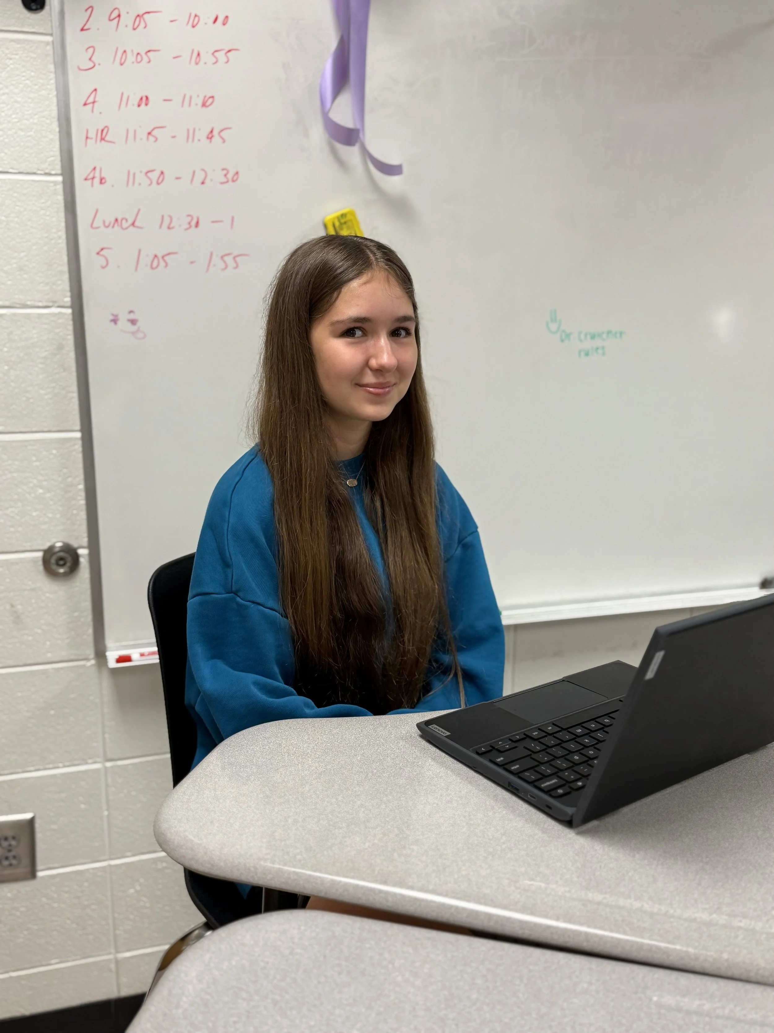 A young woman with long brown hair, wearing a blue sweatshirt, sitting at a table with a laptop, in front of a whiteboard with handwritten schedule and notes.