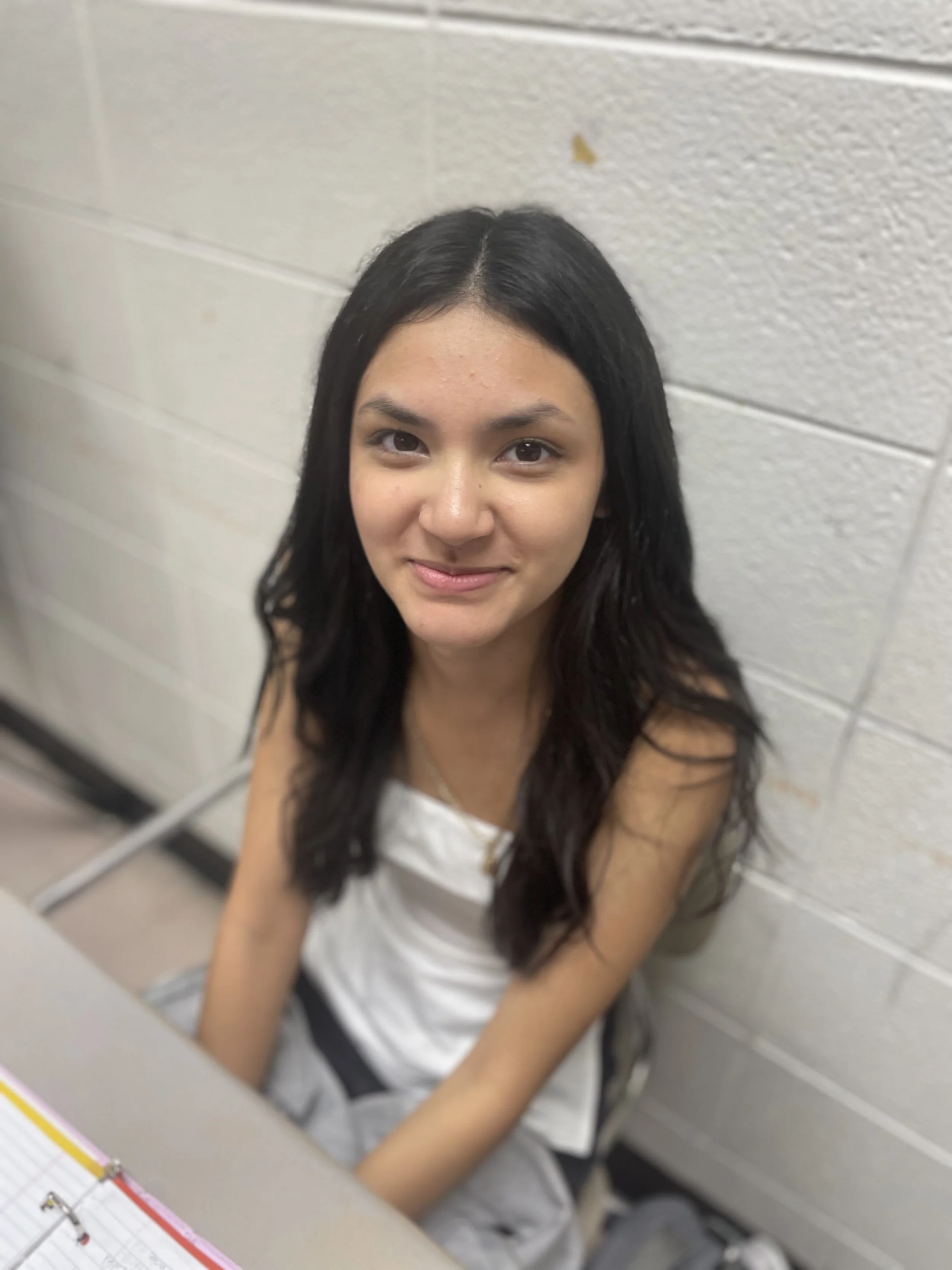 A young woman with long black hair sitting and looking directly at the camera, smiling slightly, against a white brick wall.