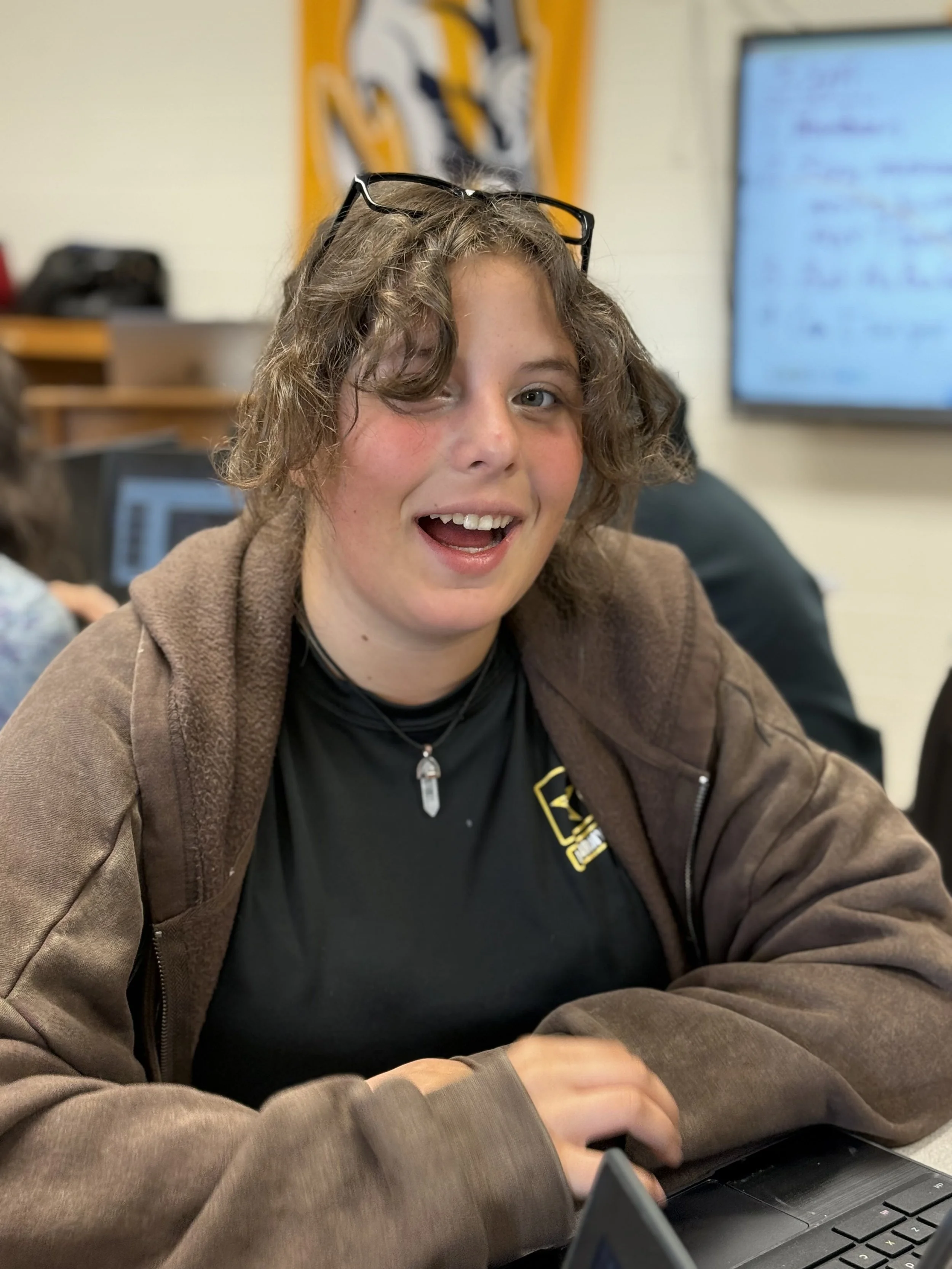 A teenage girl with curly hair, glasses resting on her head, smiling and looking at the camera in a classroom.