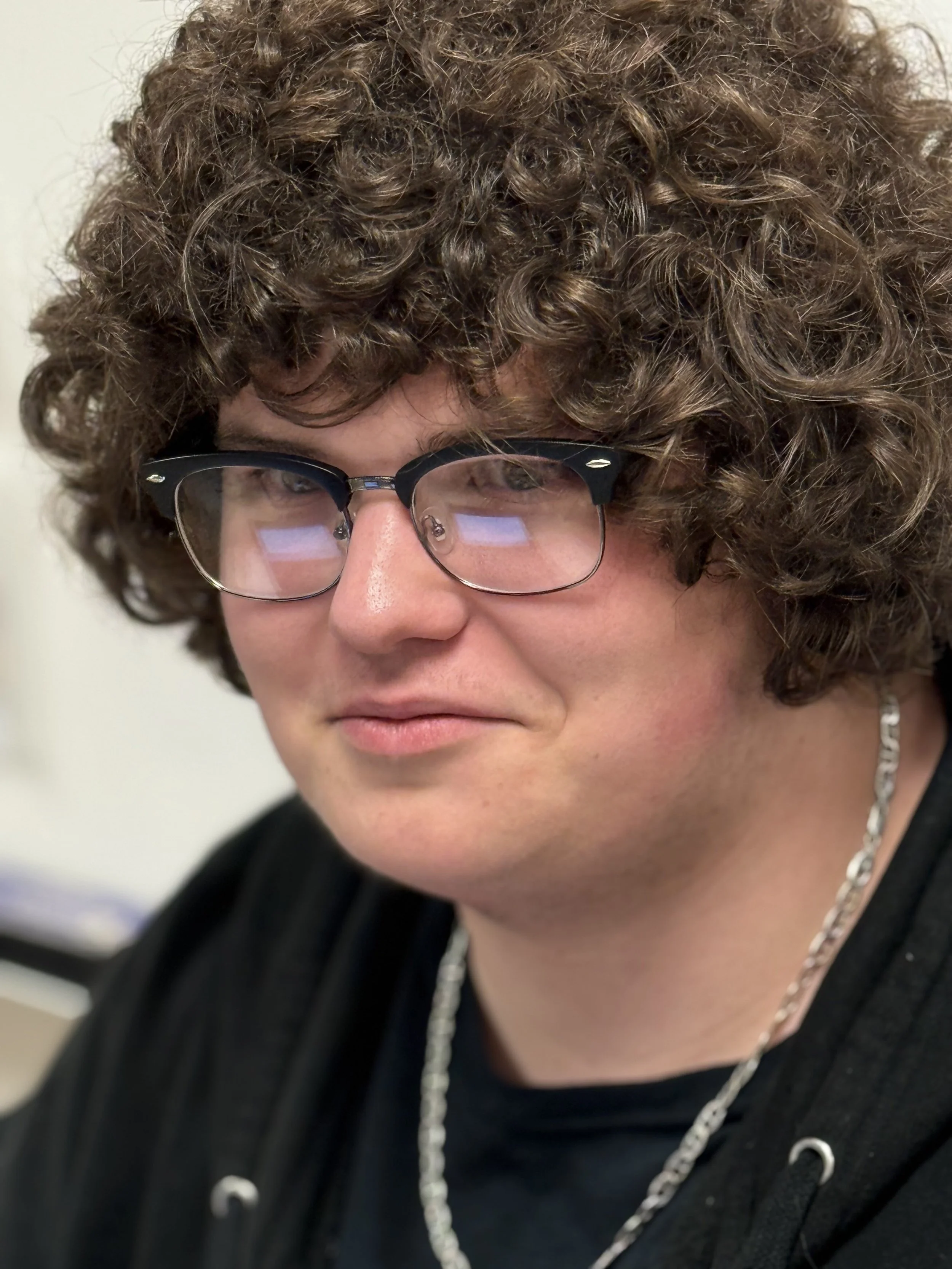 Close-up of a person with curly brown hair, wearing black-rimmed glasses, a silver chain necklace, and a black shirt.