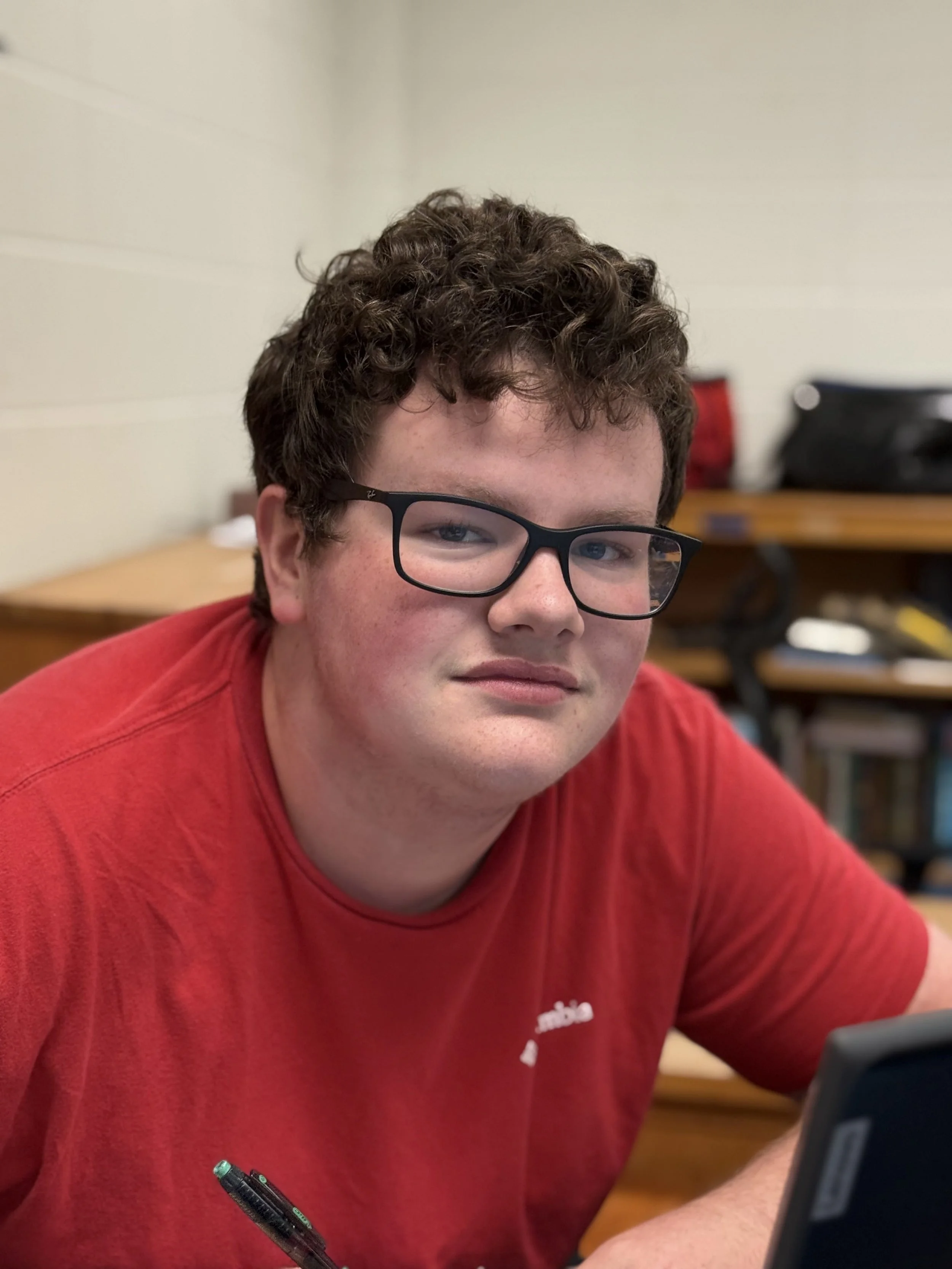 A young man with curly brown hair and glasses sitting at a desk, wearing a red shirt, with bookshelves and a desk in the background.