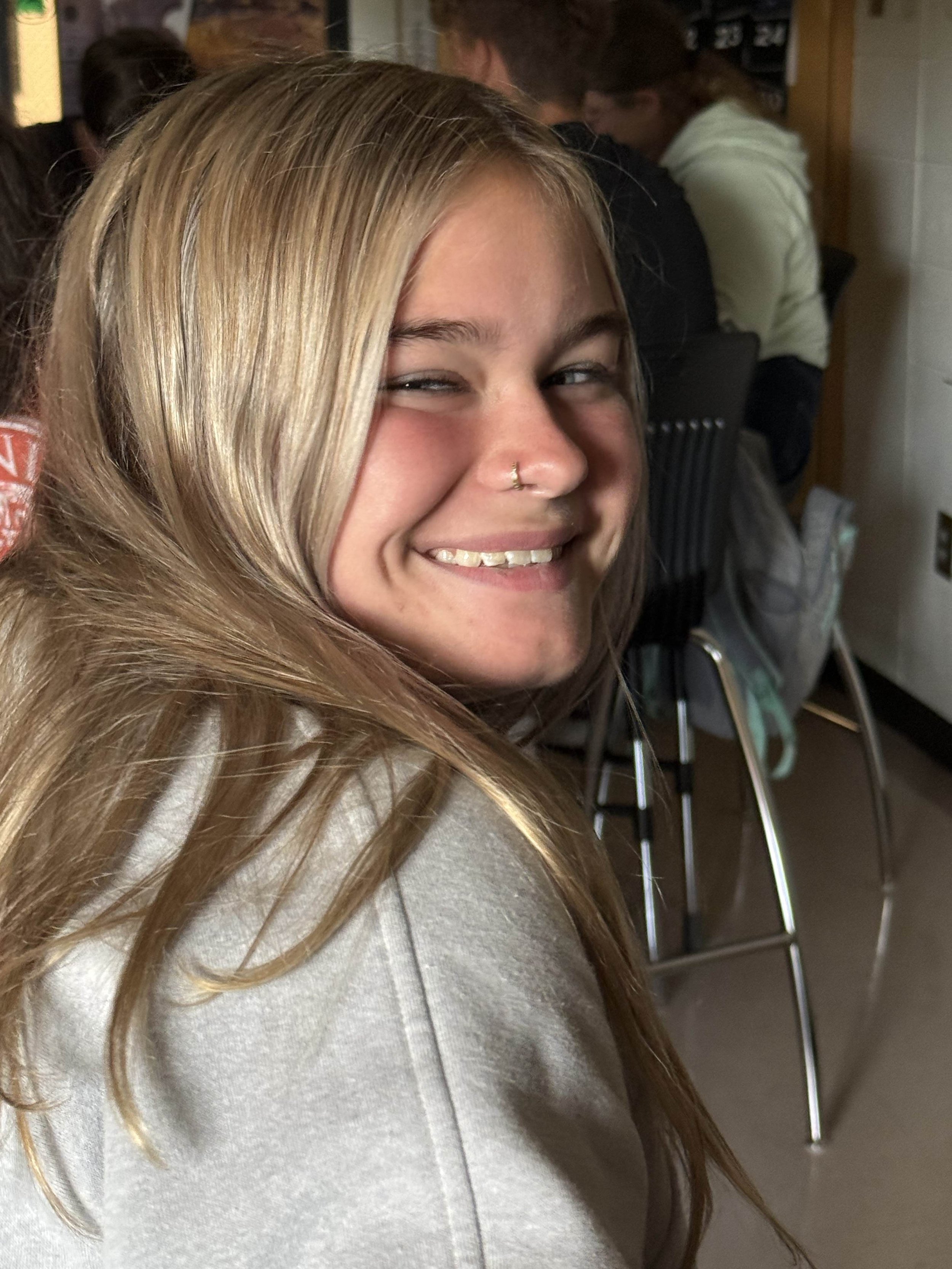 A young woman with long, blonde hair smiling at the camera, wearing a light-colored hoodie, sitting inside a crowded room.