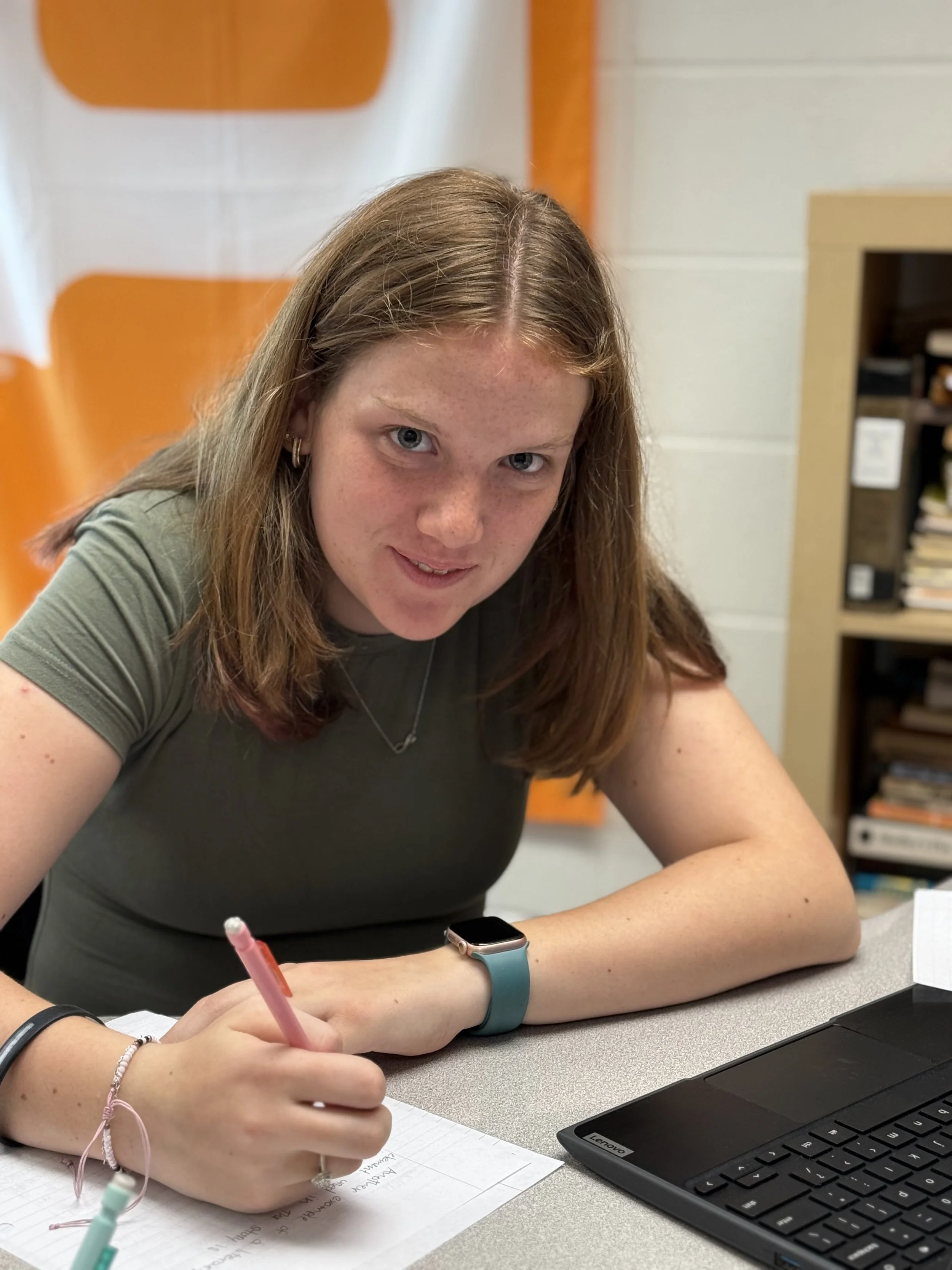 A young woman with red hair, blue eyes, and freckles sitting at a table, writing on a sheet of paper with a pink pen, with a laptop and a black smartwatch on her wrist.