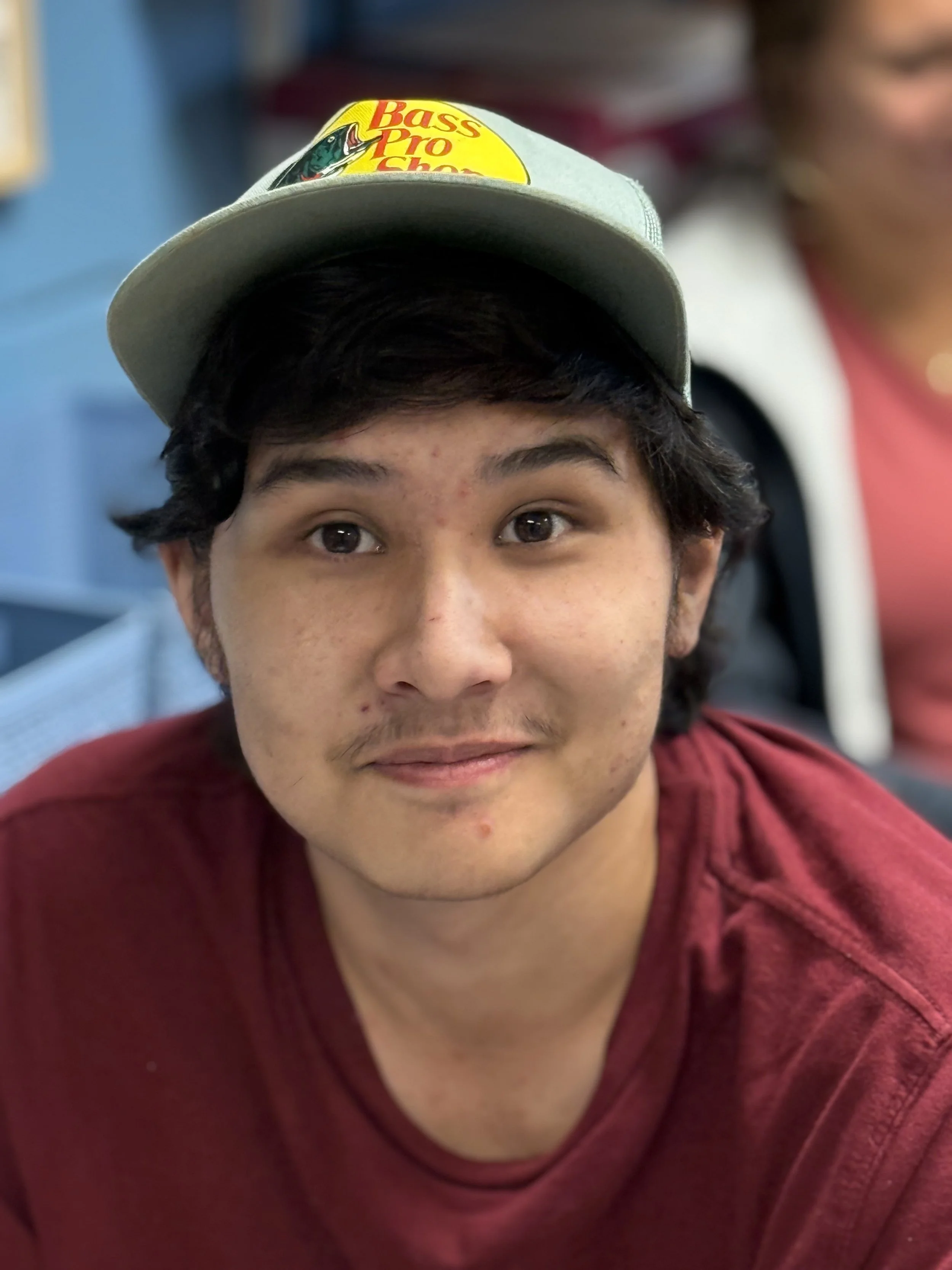 Young man with dark hair, eyebrows, and slight facial hair wearing a gray baseball cap with a colorful Bass Pro Shops logo. He is smiling slightly and wearing a red shirt, sitting indoors with a blurred background of blue seats and a woman in a pink shirt.
