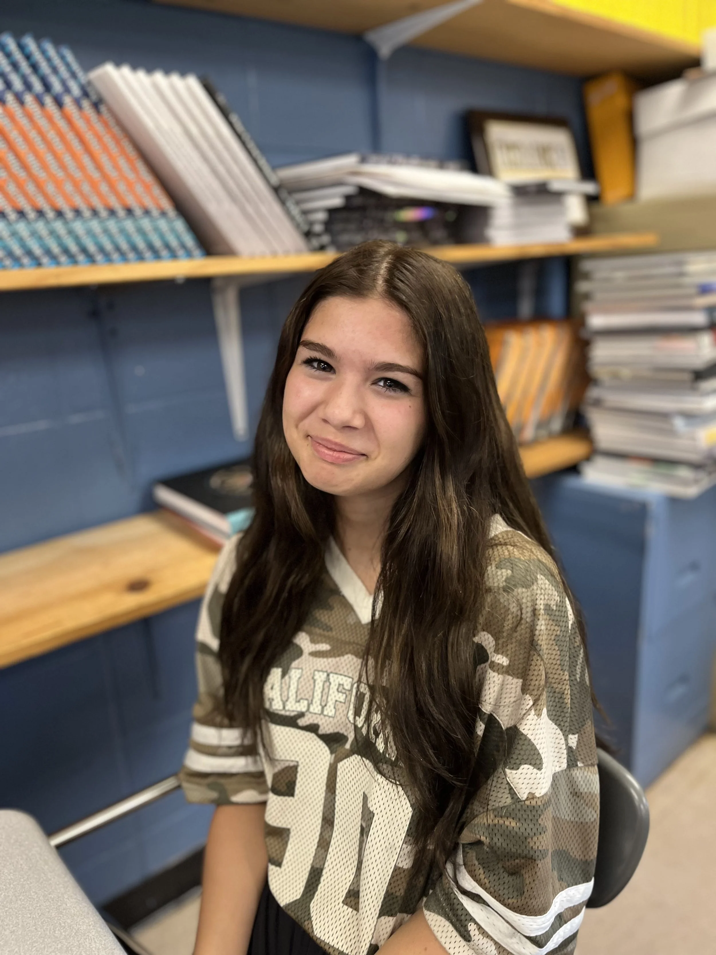 A girl with glasses and a pink Nike t-shirt is sitting at a desk in a classroom, smiling and looking at the camera. She is wearing an Apple Watch and a necklace, with a purple notebook in front of her. Other students are visible in the background.