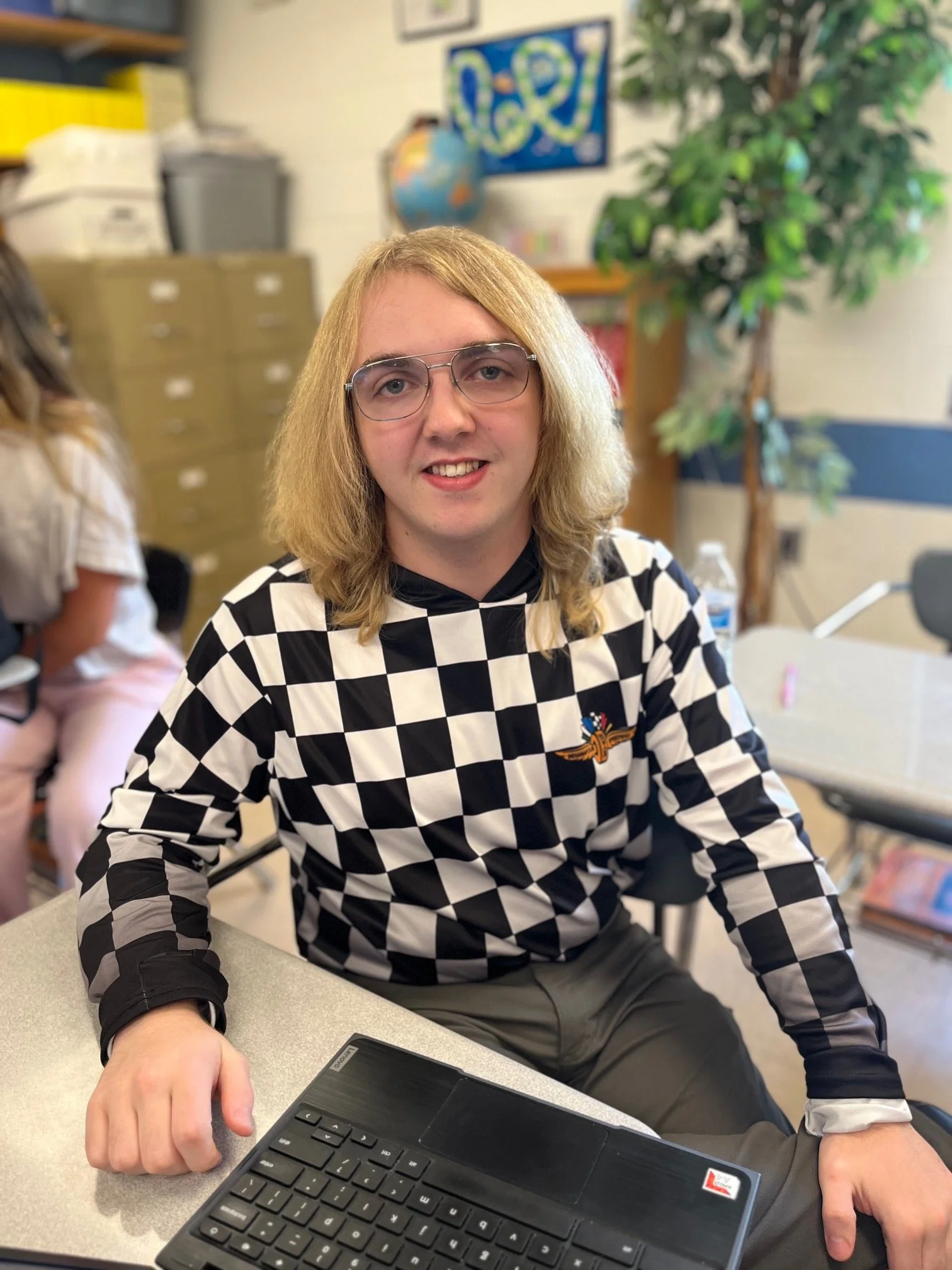 Young man with shoulder-length blond hair, glasses, wearing a black polo shirt with a 'Panther Robotics' logo, smiling at the camera in an indoor setting.