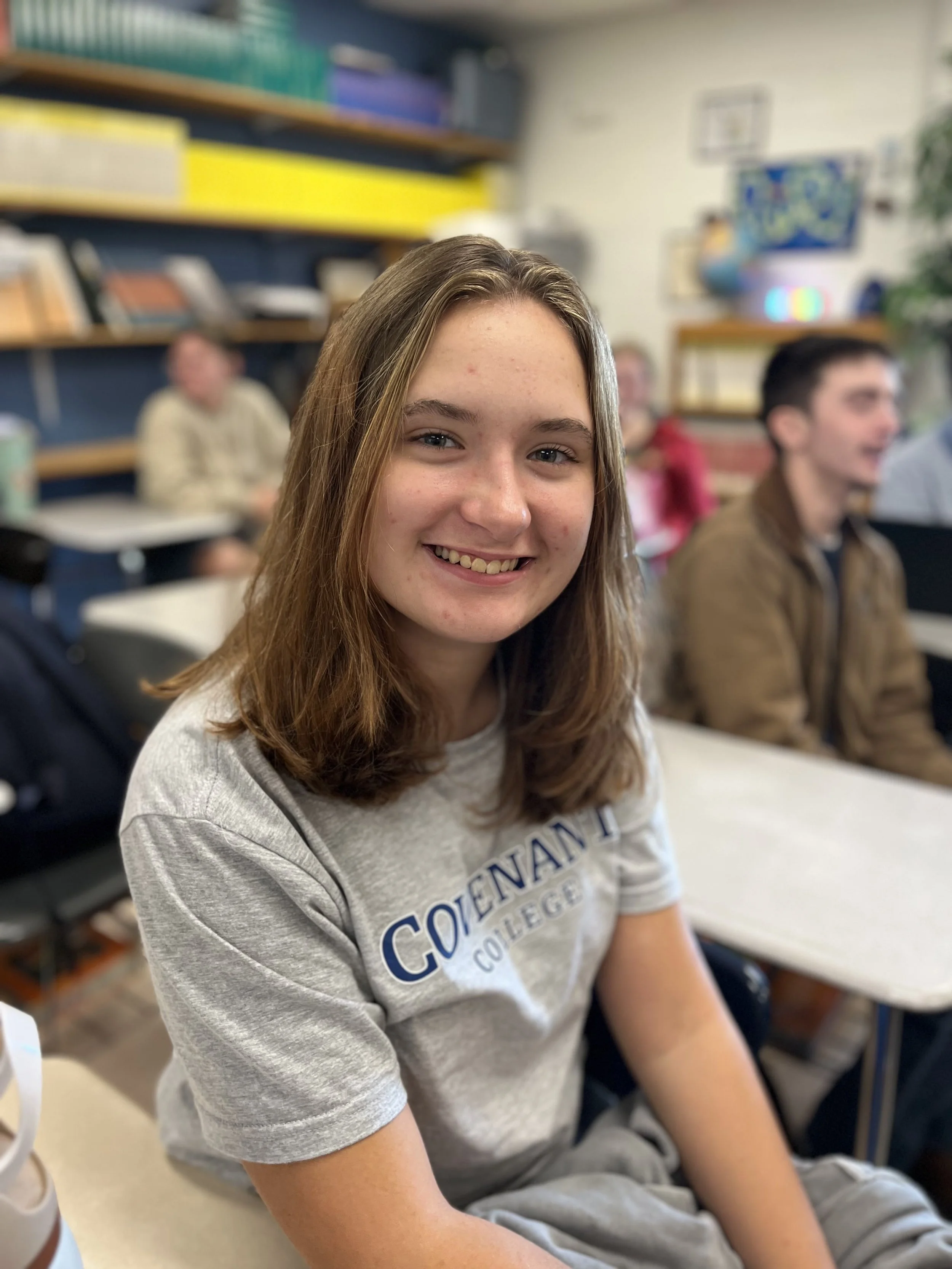 A young girl with brown hair smiling at the camera, sitting at a desk with a notebook and a pen. In the background, there are posters and other students in a classroom.