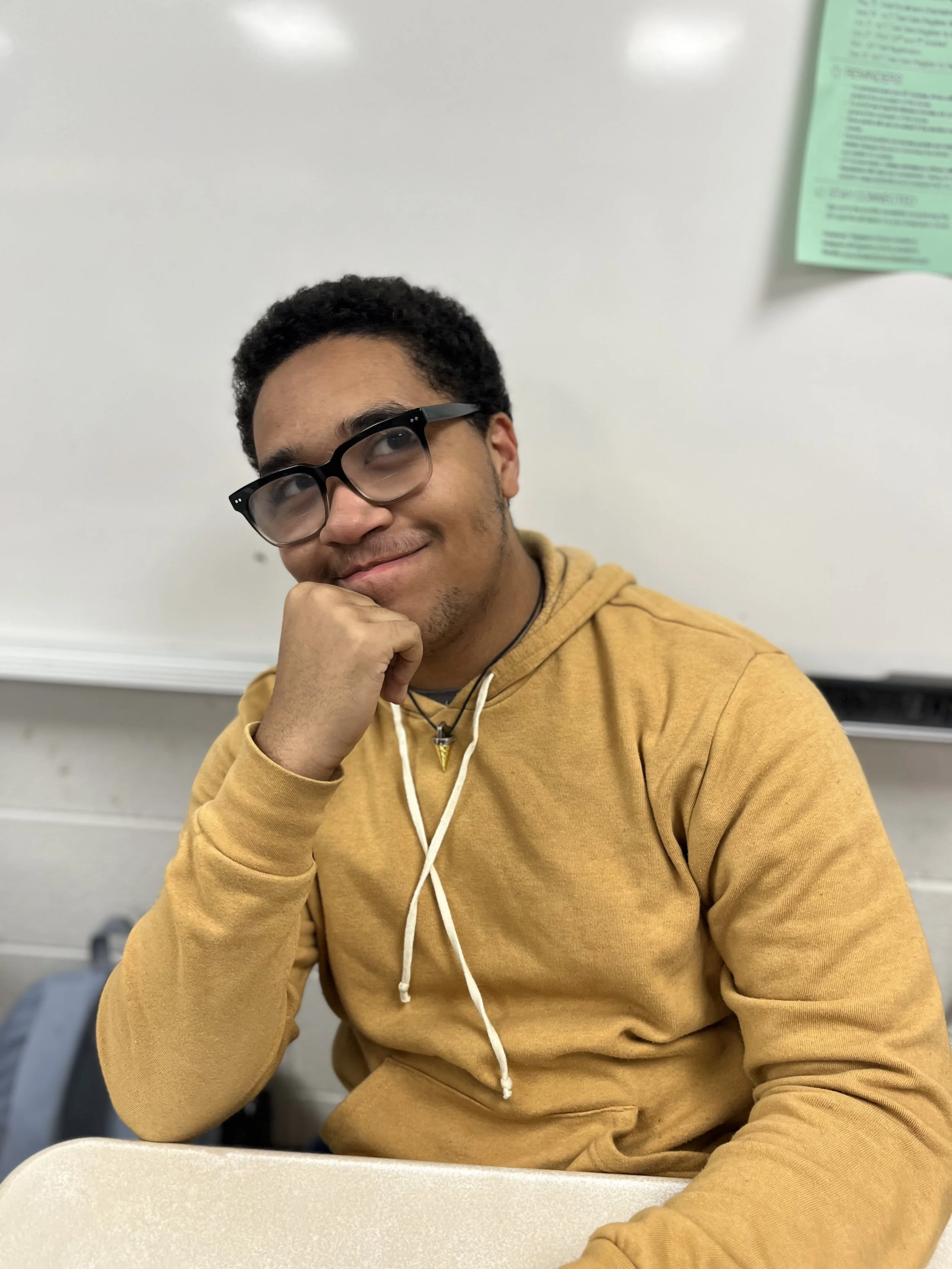 A young man with curly dark hair, wearing black glasses and a gray hoodie, smiling and looking over his shoulder in a classroom setting.