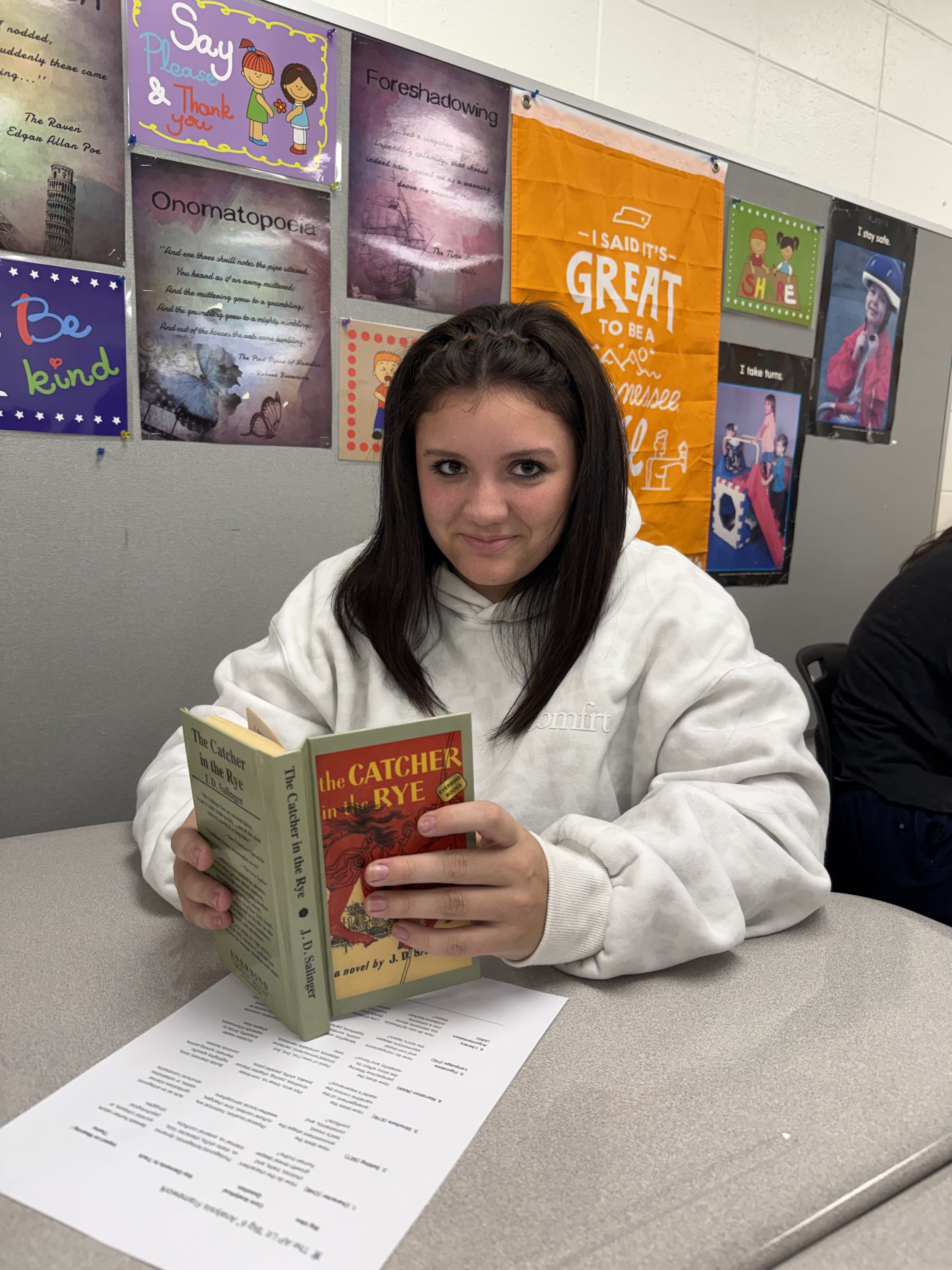 A girl with braided hair and a beige T-shirt smiling at the camera in a classroom with other students working at their desks.