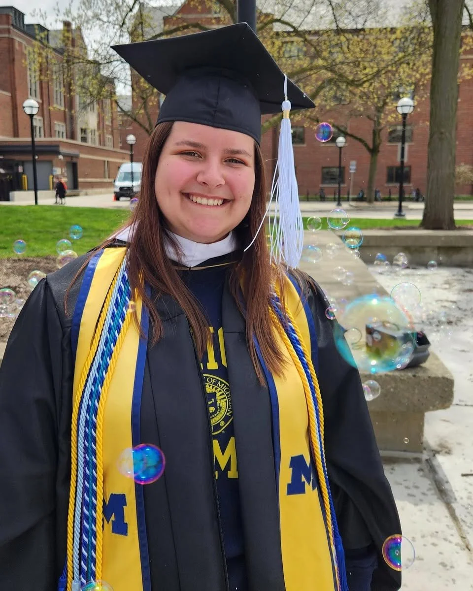 A young woman in graduation attire smiling outdoors, wearing a black cap with a white tassel, a black gown, and a yellow and blue stole with cords. There are soap bubbles floating around her with a school building and trees in the background.