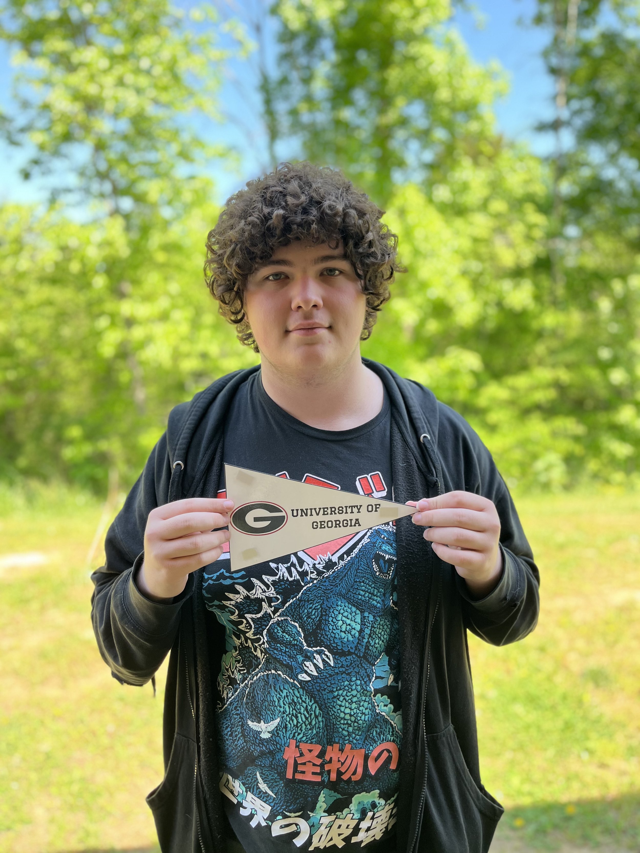 Close-up of a person with curly brown hair, wearing black-rimmed glasses, a silver chain necklace, and a black shirt.