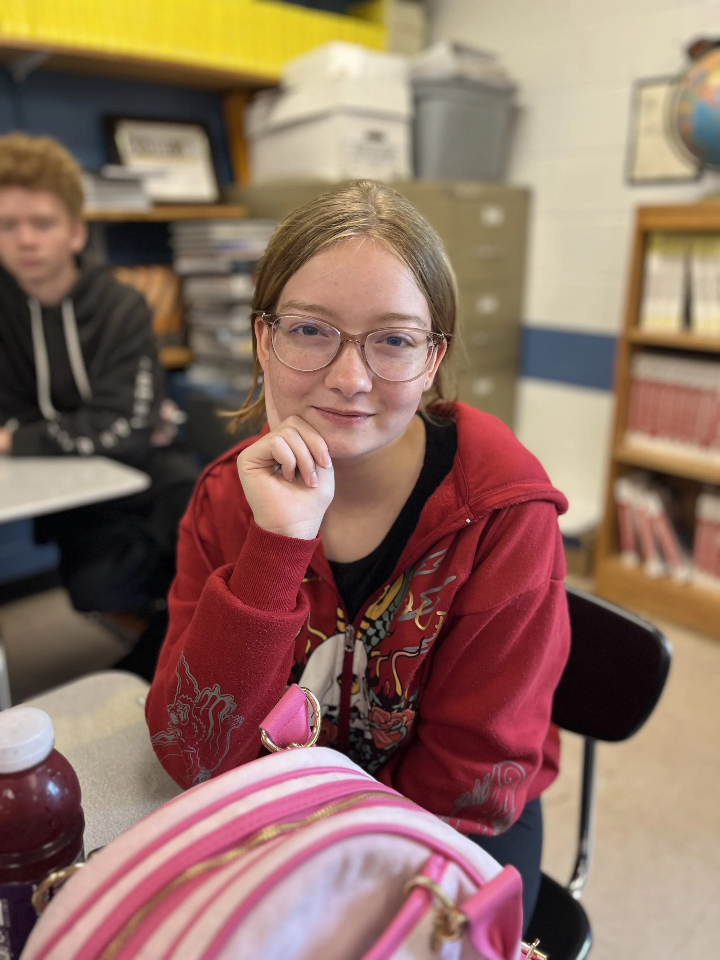 A girl with glasses smiling at the camera, sitting at a desk in a classroom, with a large screen and other students in the background.