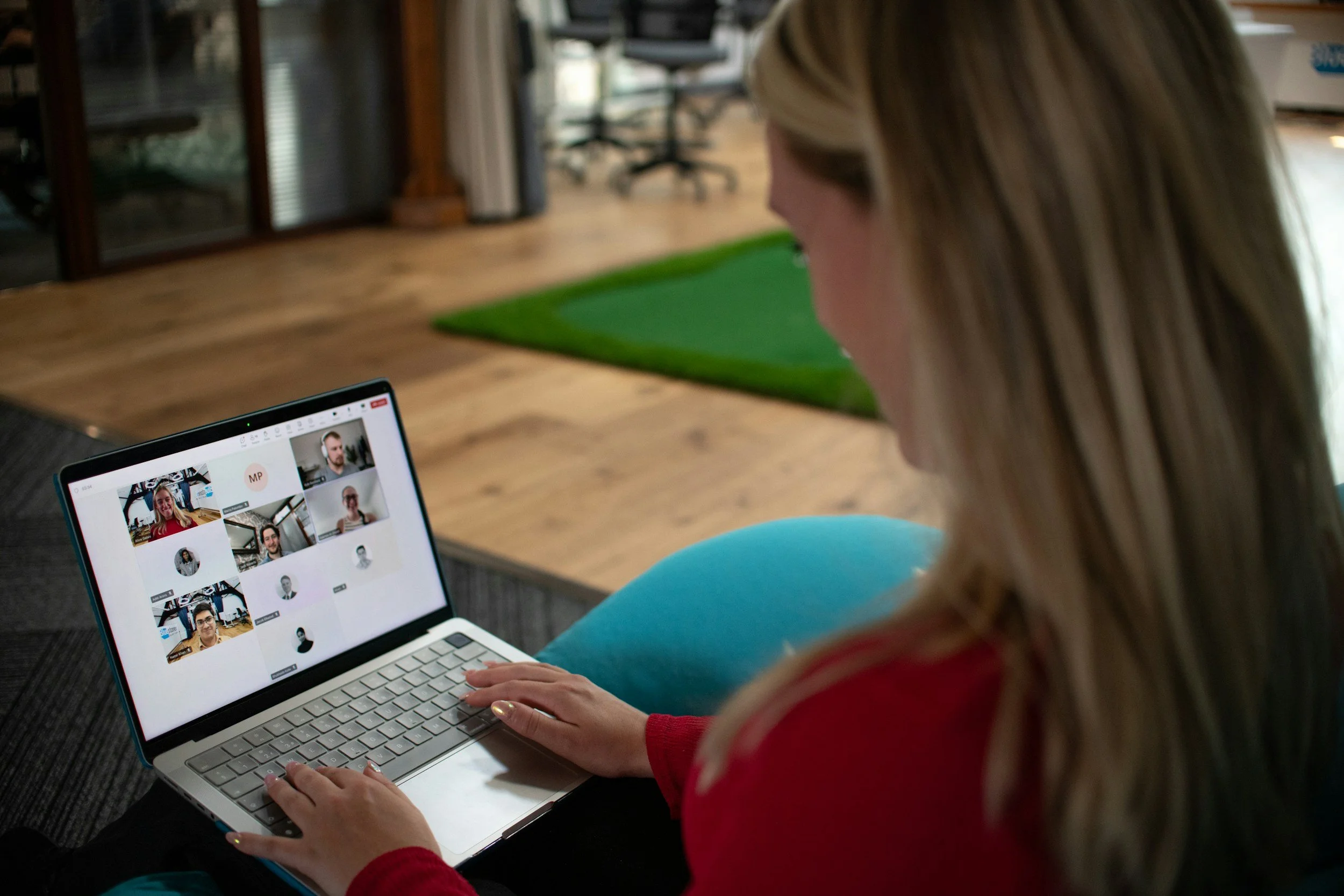 A woman is sitting on the floor using a laptop for a video conference call with multiple participants.