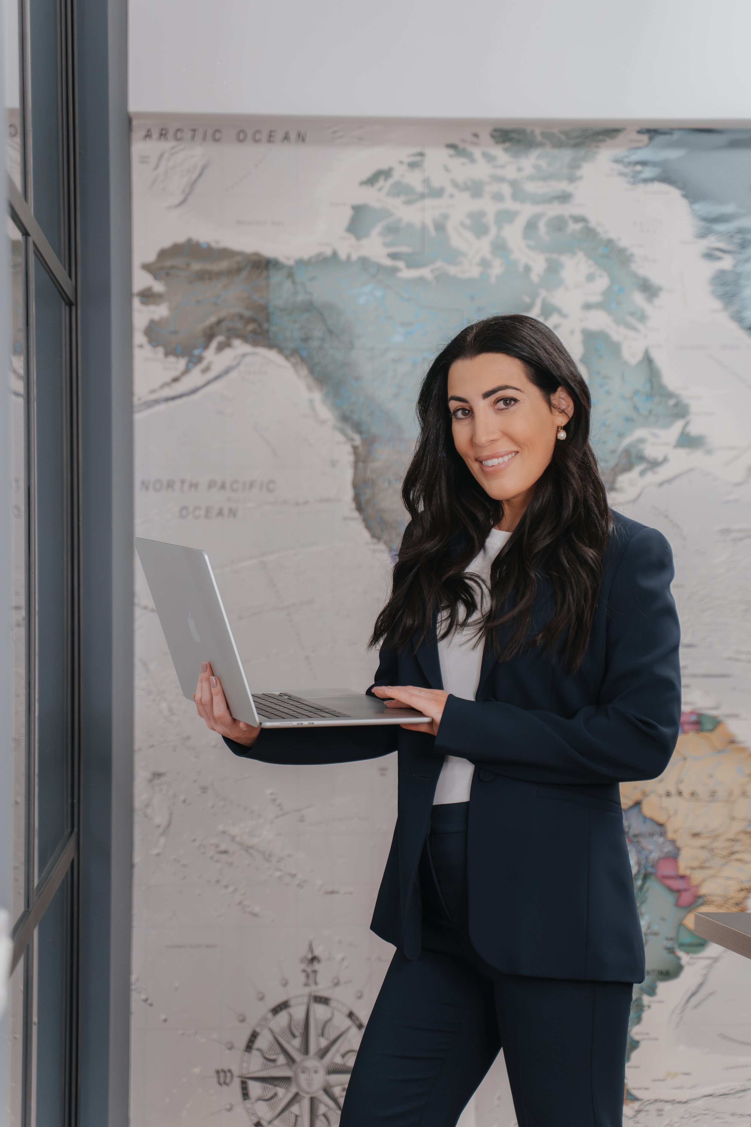 A woman in a business suit holding a laptop, standing in front of a world map and a clock, in an office environment.