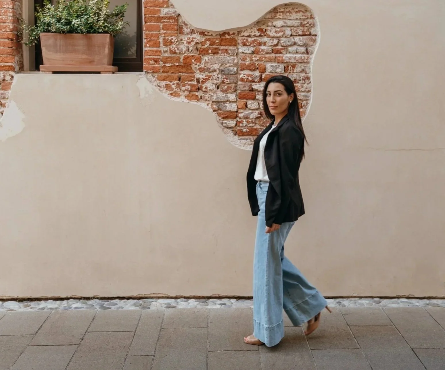 Woman standing in front of a wall with a section of exposed brick, wearing a black blazer, white blouse, wide-leg jeans, and sandals.
