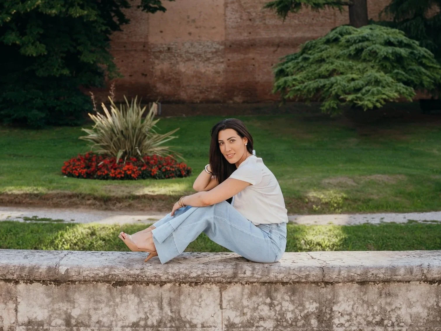 A young woman sitting on a stone ledge in a park with greenery, flowers, and a brick wall in the background.