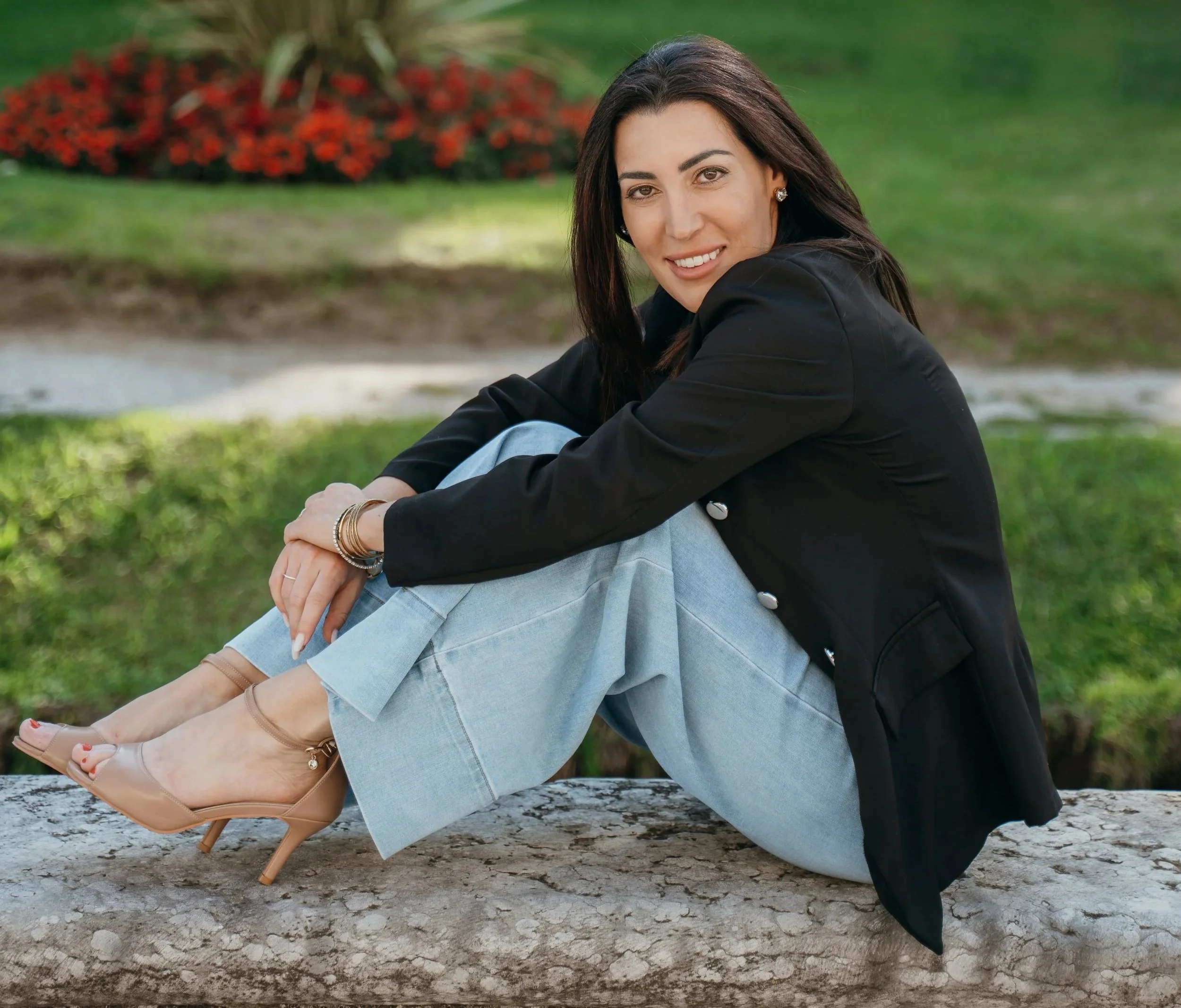 A woman with dark hair, wearing a black blazer, light blue jeans, and nude high heels, sitting on a log outdoors with a grassy background and red flowers.