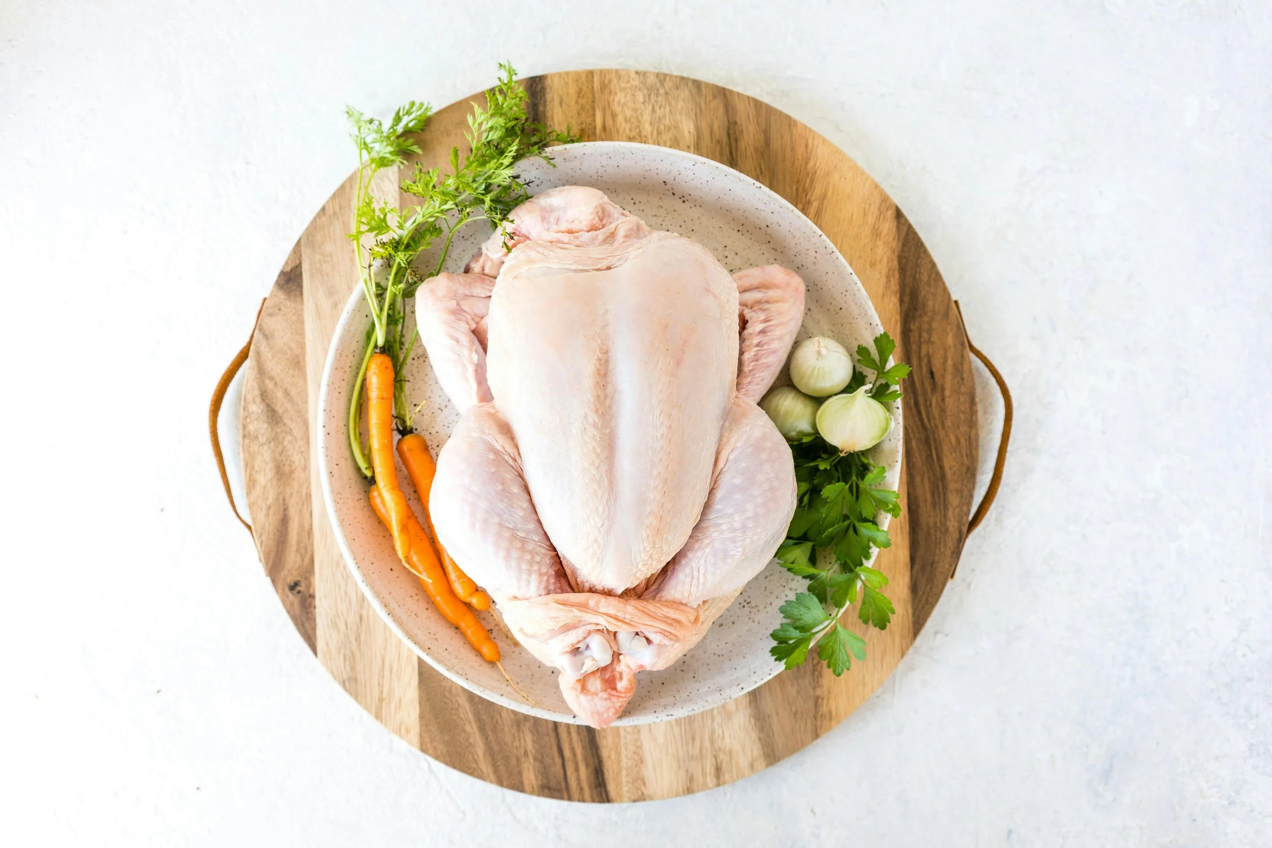 Raw whole chicken on a platter with garlic, carrots, parsley, and cilantro, on a wooden tray.