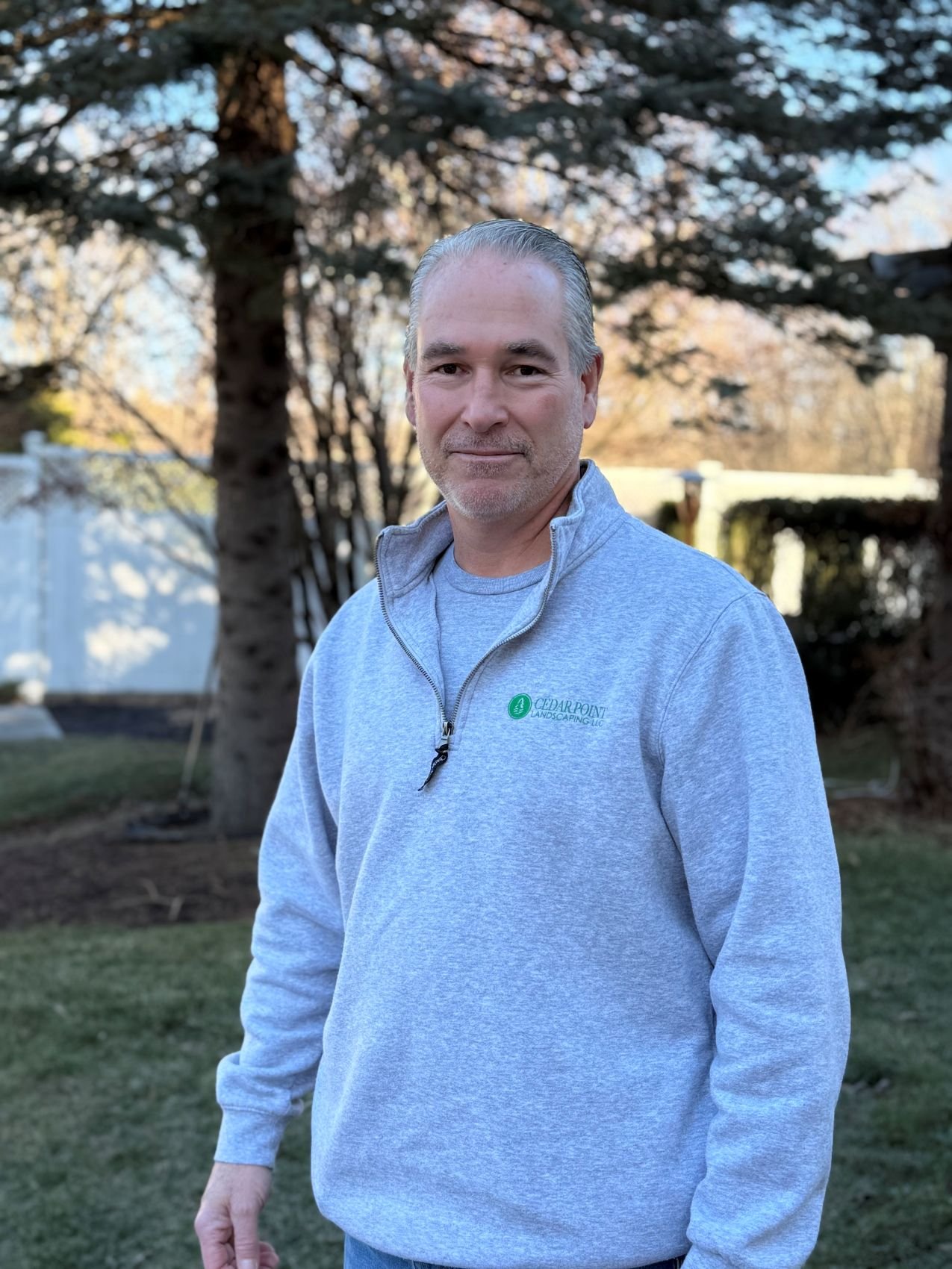 A middle-aged man with gray hair and a beard standing outdoors in a backyard with trees and a white fence, wearing a light gray zip-up sweatshirt with a company logo.