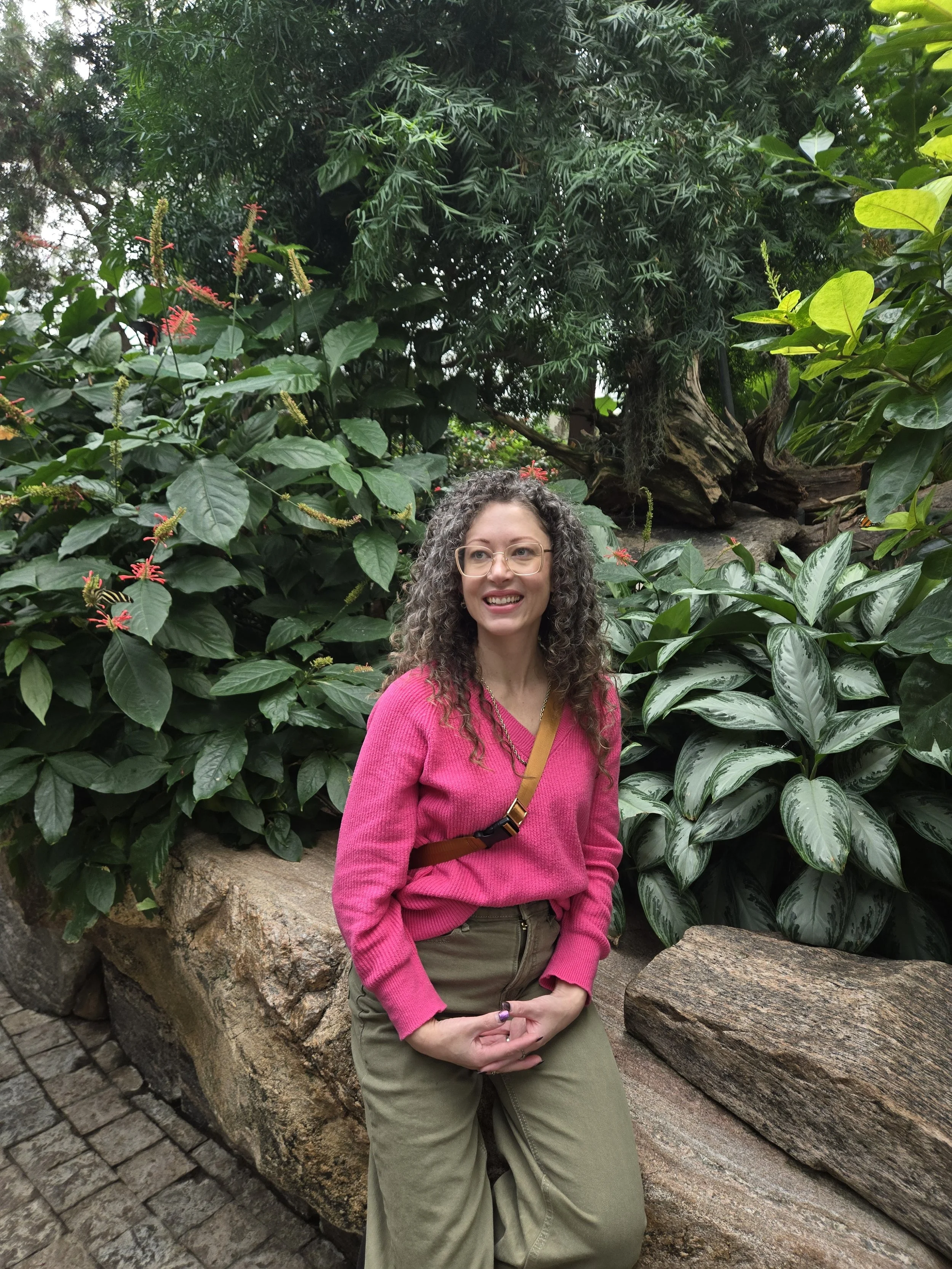 Woman with curly hair wearing a pink sweater and khaki pants, sitting on a large rock surrounded by lush green tropical plants.