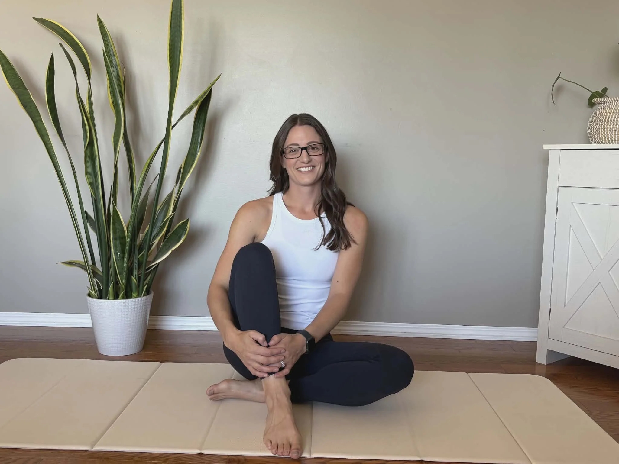 A woman with long brown hair, glasses, white tank top, and black pants sitting on a beige yoga mat on hardwood floor, smiling, with a large potted plant and a white cabinet in the background.