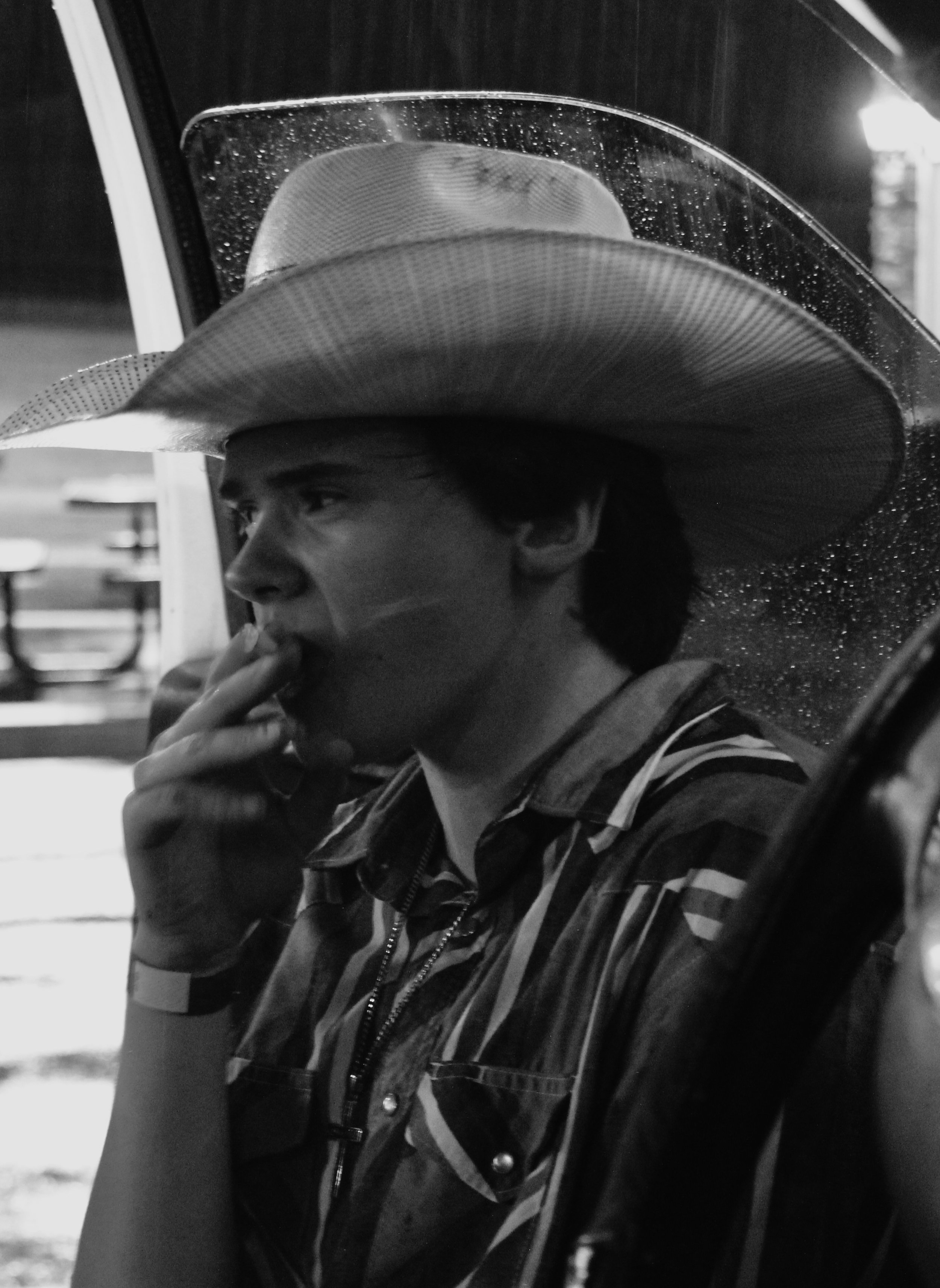A black and white photo of a man wearing a large wide-brimmed hat, sitting outdoors at night, holding a cigarette near his mouth.