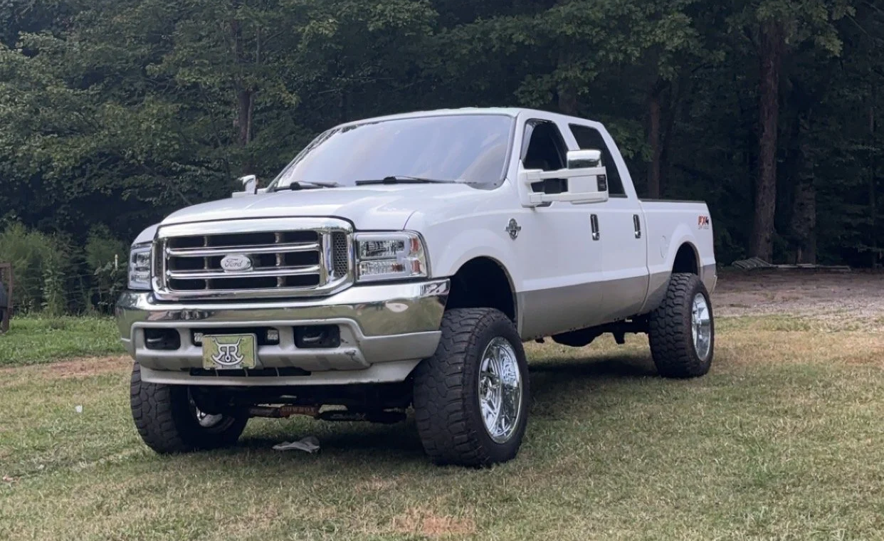 A white lifted Ford pickup truck with large off-road tires parked on grass with trees in the background.
