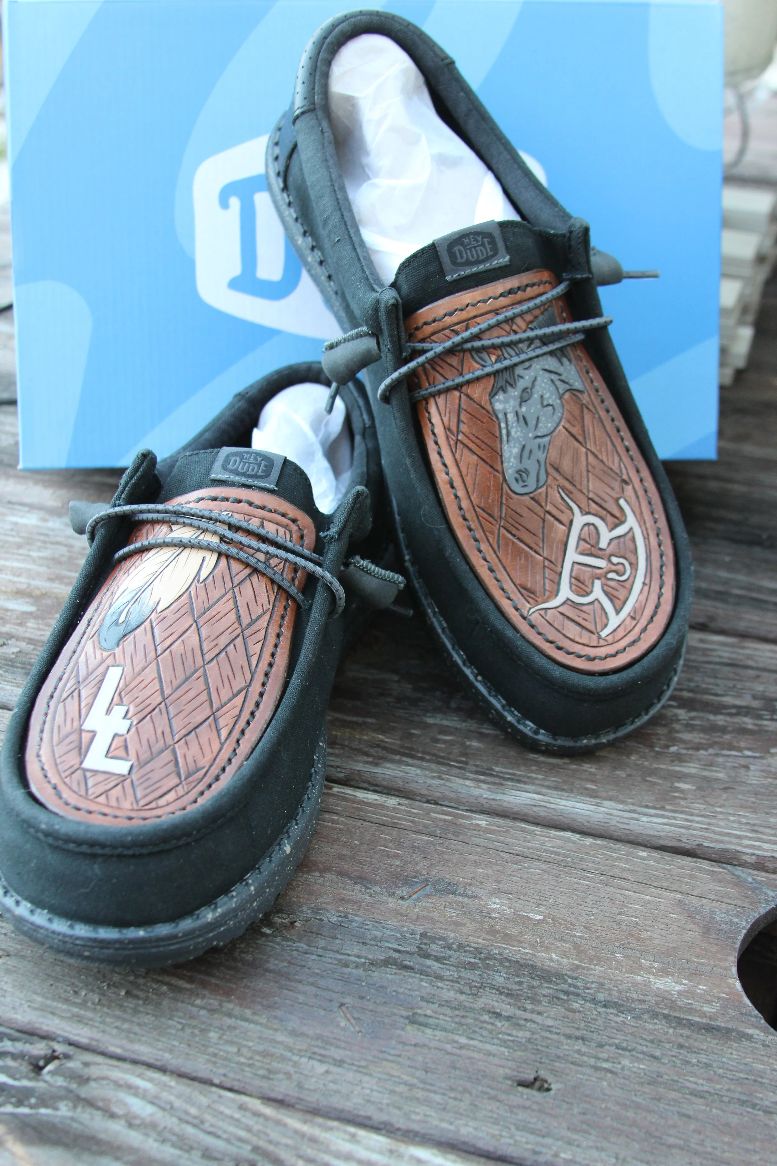 Pair of black slip-on shoes with brown leather patches featuring a horse head and feather designs, placed on a wooden surface with a blue box in the background.