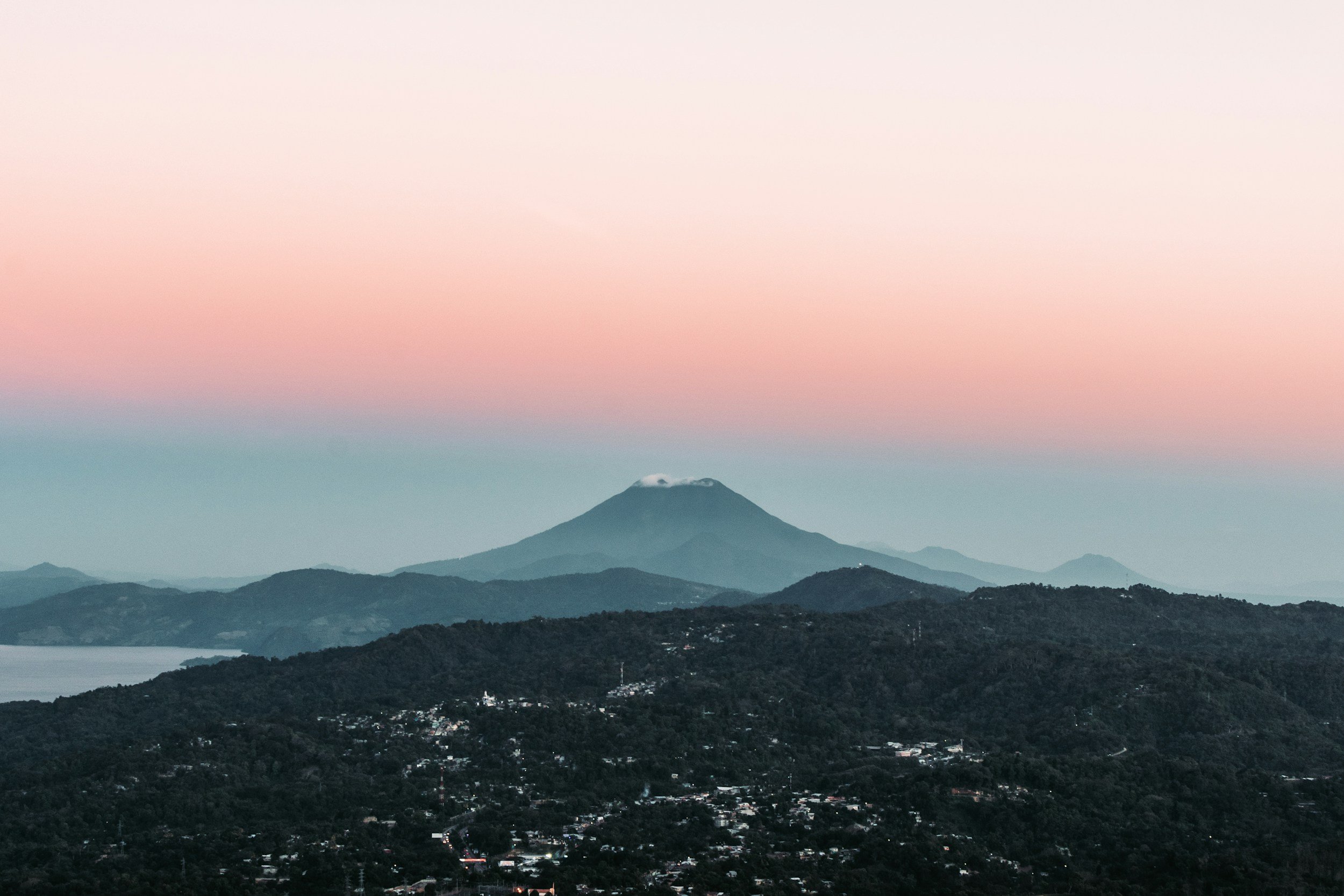 Vista de un volcán con una cresta blanca, rodeado de colinas verdes y un cielo con tonos rosados y azules