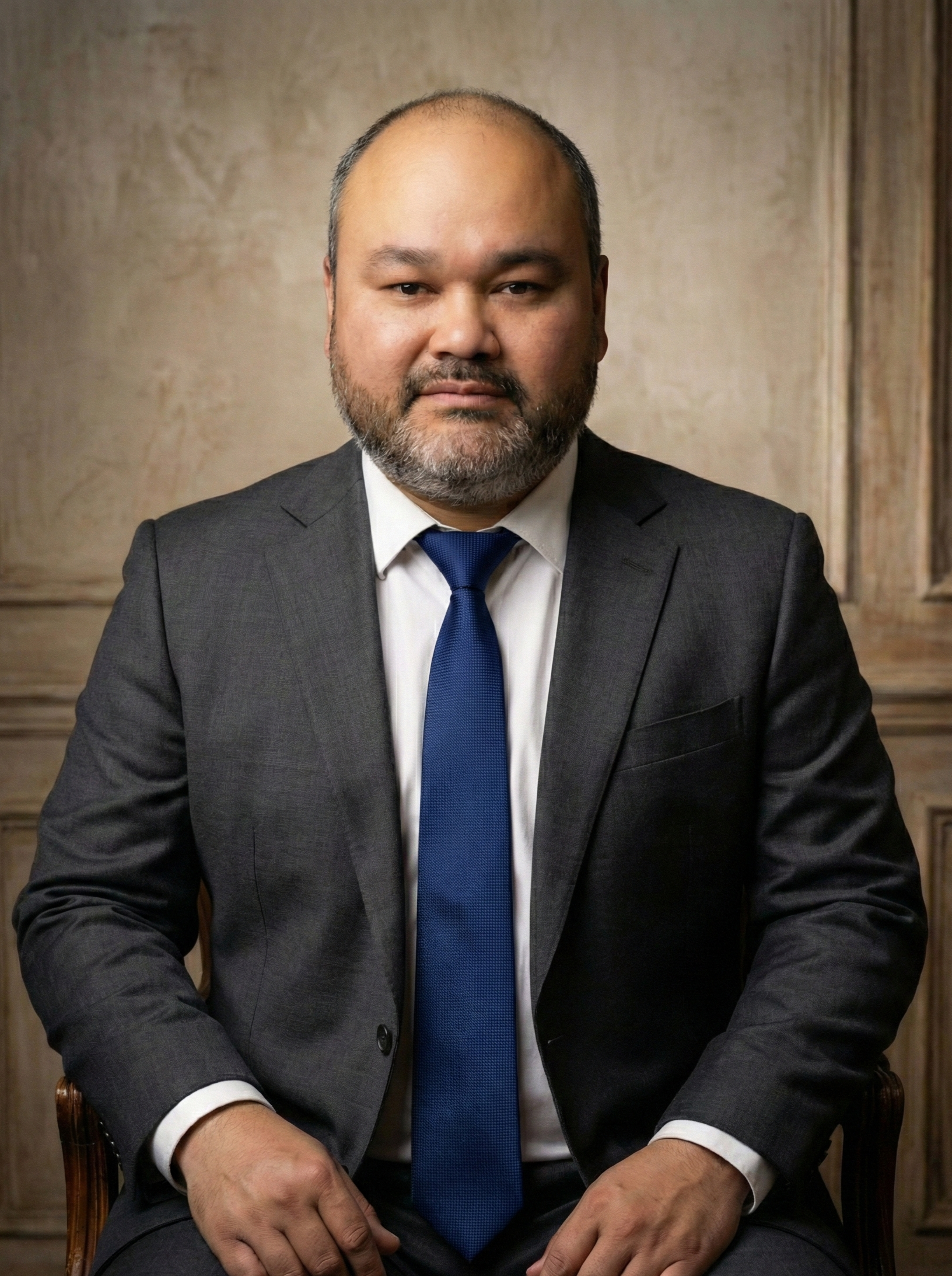 Hombre con barba y barba, usando un traje gris y una corbata azul, sentado en una silla, en un fondo de pared de madera