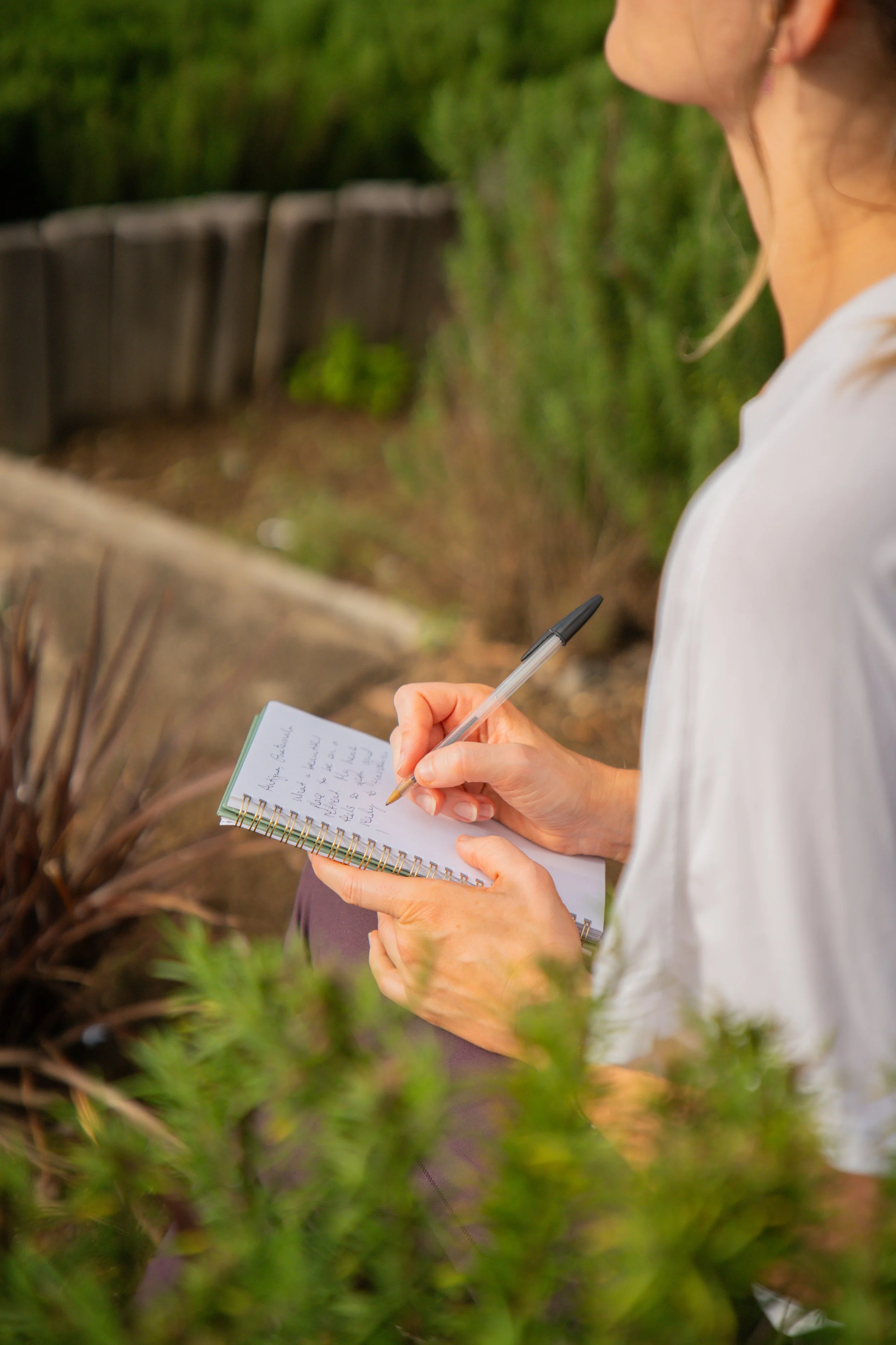 Woman sitting outdoors, writing in a spiral notebook with a pen, surrounded by greenery.
