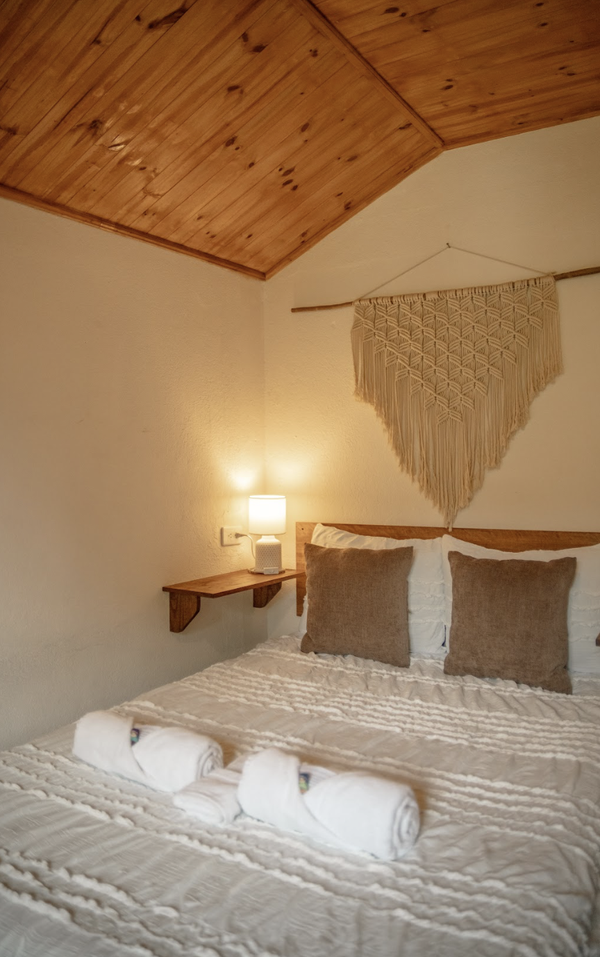 A cozy bedroom with a wooden ceiling, beige walls, and a bed with white bedding and three rolled towels. There are two brown pillows on the bed, a small wooden nightstand with a lamp, and a beige macrame wall hanging above the bed.