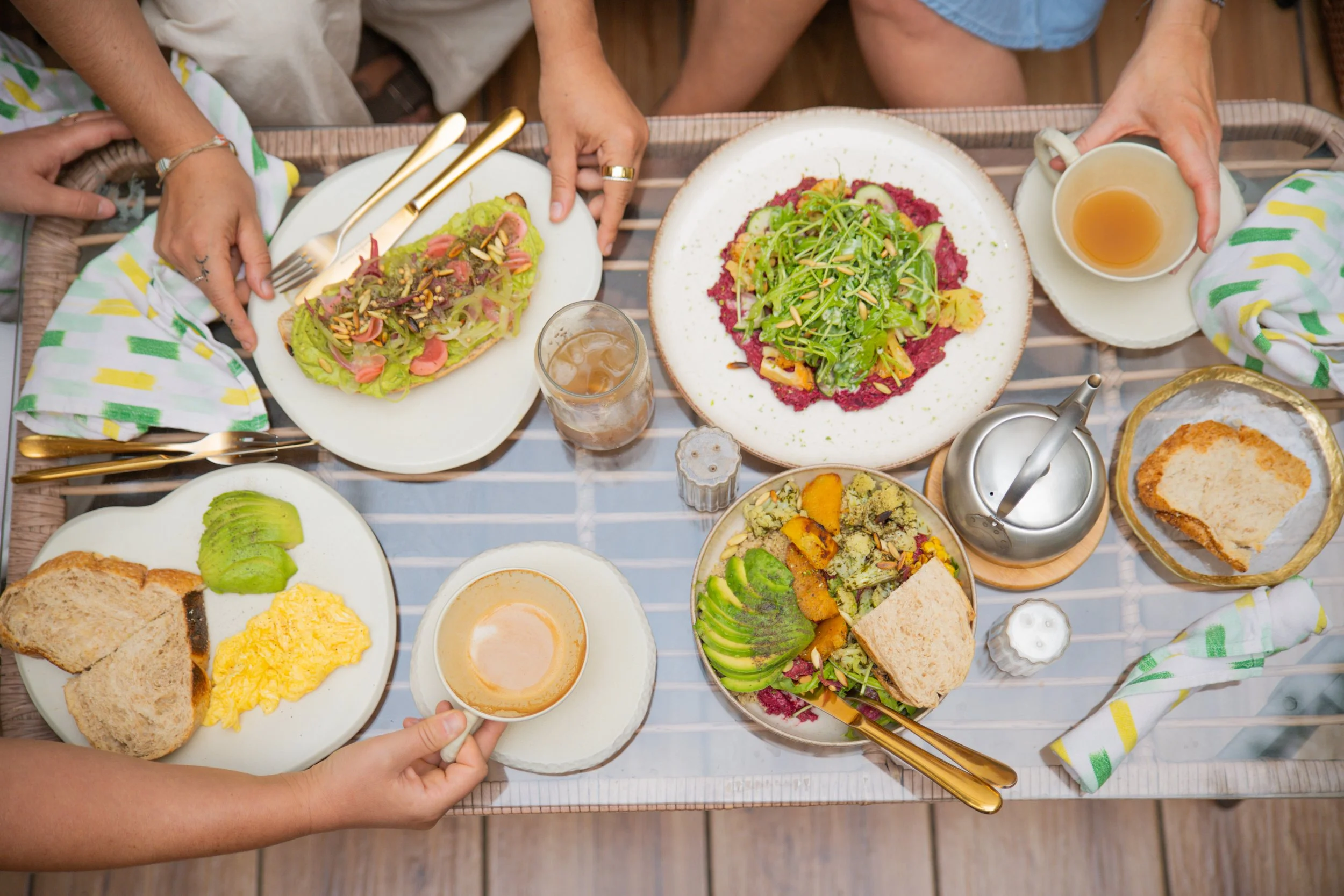 Overhead view of a breakfast or brunch table with various dishes including sliced avocado, scrambled eggs, toast, salads with greens and vegetables, a cup of tea, a glass of iced coffee, and a small teapot.