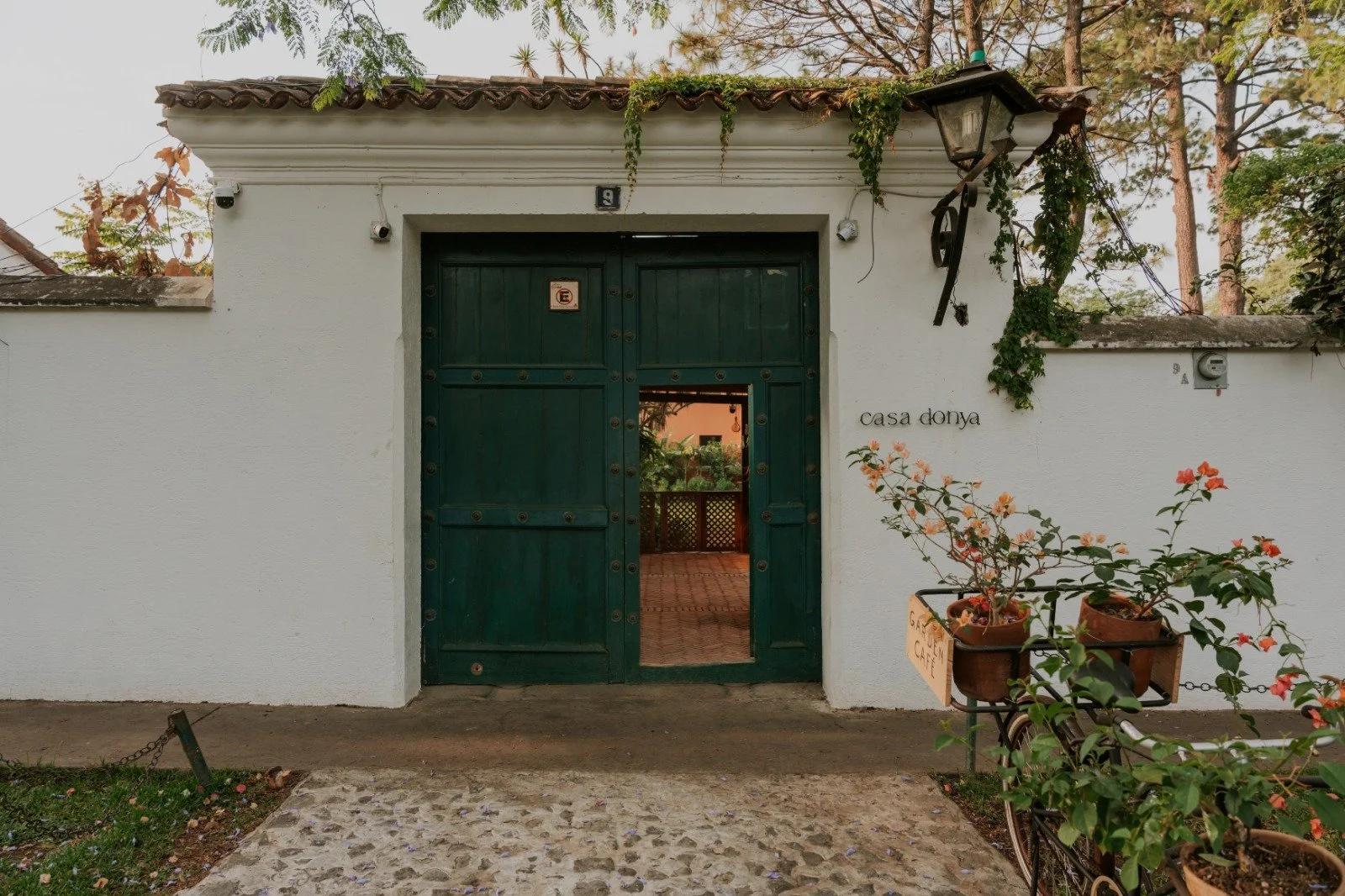 White wall with a dark green double door, a small mirror above, two security cameras, a vintage wall-mounted lantern, and potted plants nearby. The words "casa donya" are painted on the wall to the right of the door.