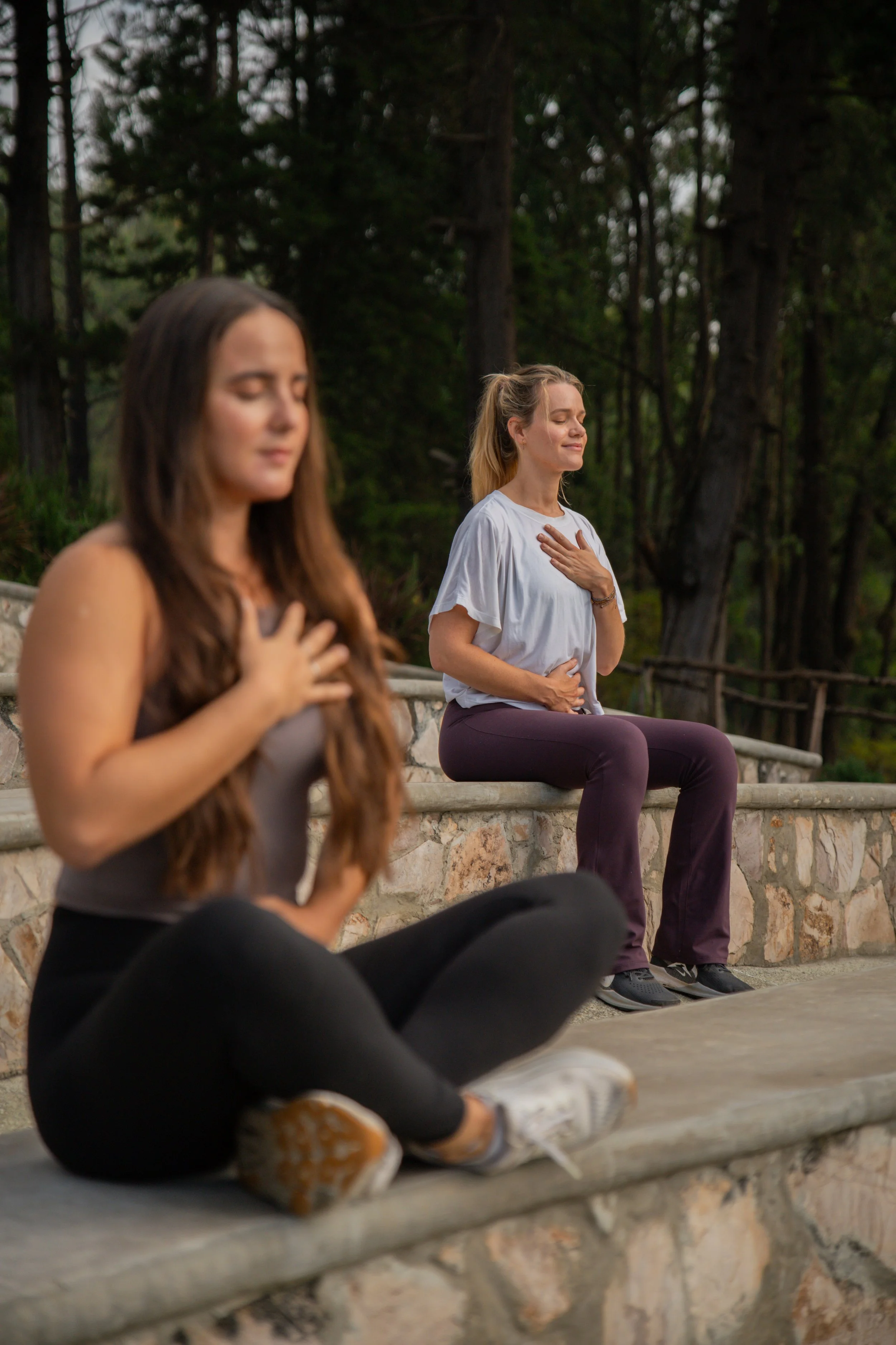 Two women practicing meditation outdoors, sitting cross-legged on a stone bench, with hands on their chests and stomachs, eyes closed, surrounded by trees.