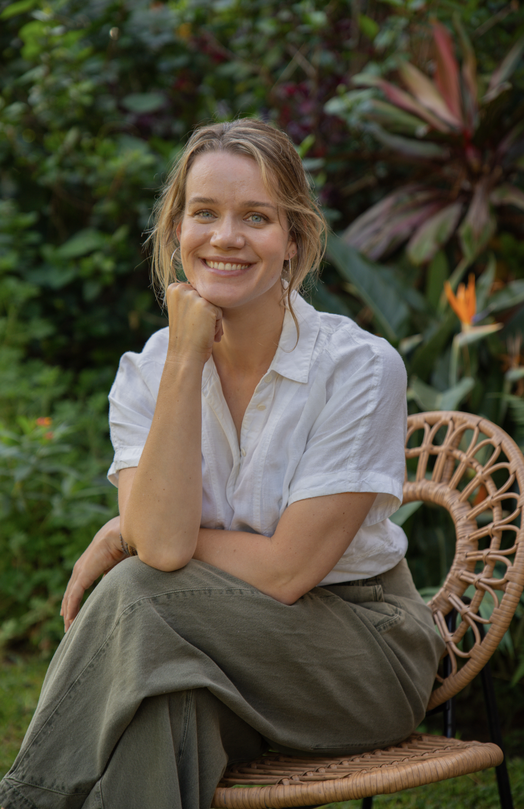 A woman sitting outdoors on a wicker chair, smiling, with lush green plants and large leaves in the background.
