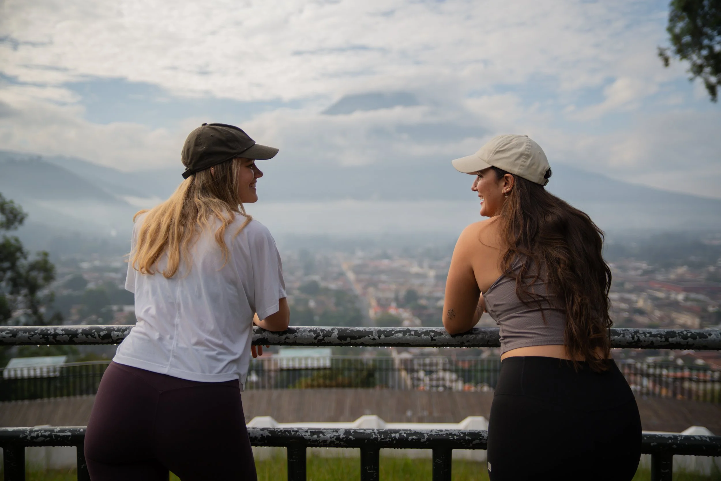 Two women standing at a railing overlooking a cityscape with mountains and clouds in the background, talking and smiling at each other on a cloudy day.